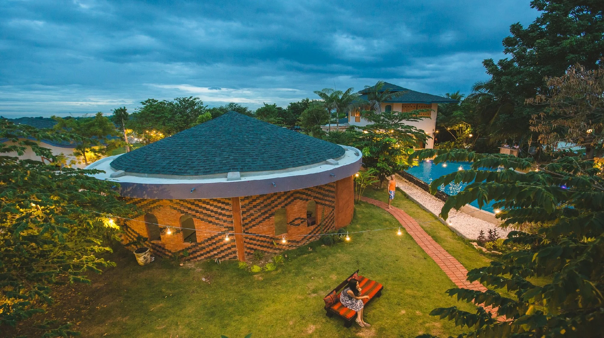 An outdoor pool area at dusk with a building with decorative brickwork, a brick walkway, a person walking by the pool, and a woman sitting on a bench under trees, with clouds in the sky.