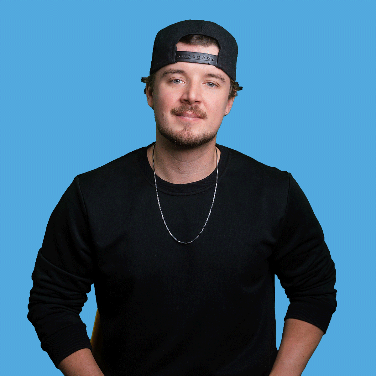 A young man with a goatee and mustache, wearing a black snapback hat, black shirt, silver chain necklace, against a blue background.