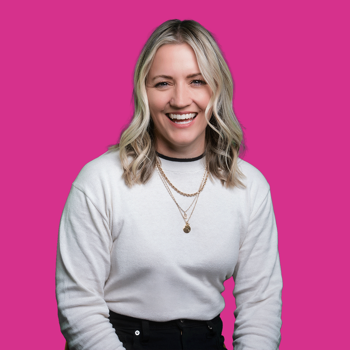 A woman smiling with blonde wavy hair, wearing a white sweatshirt with black trim, layered gold necklaces, and standing against a pink background.