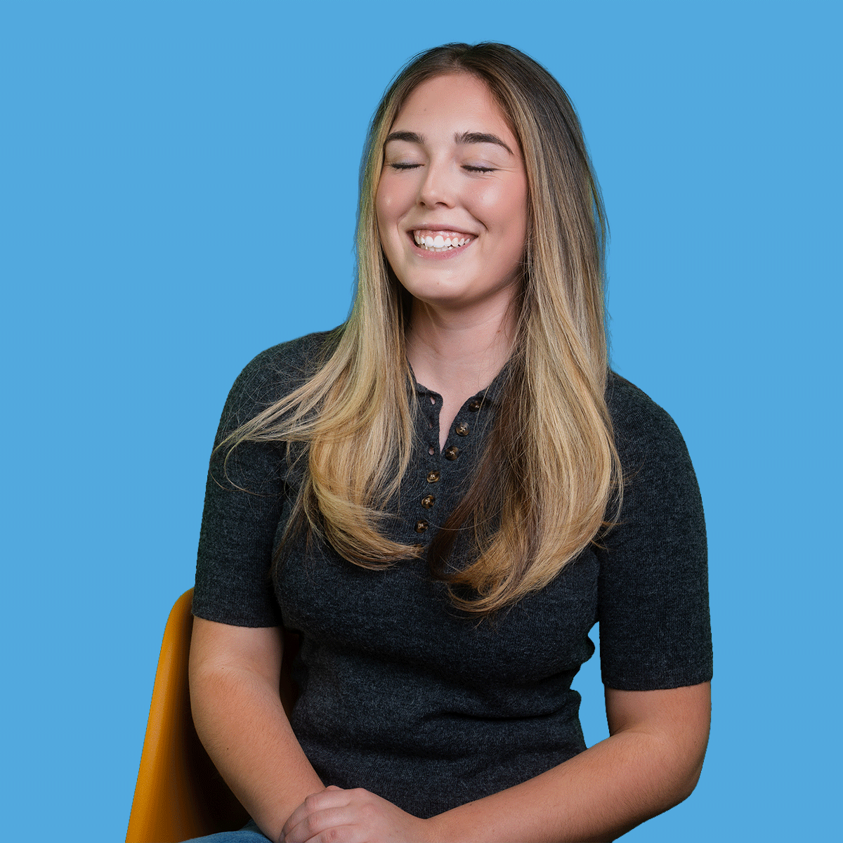 A smiling young woman with long, wavy hair, closed eyes, wearing a dark gray collared shirt, seated against a blue background.