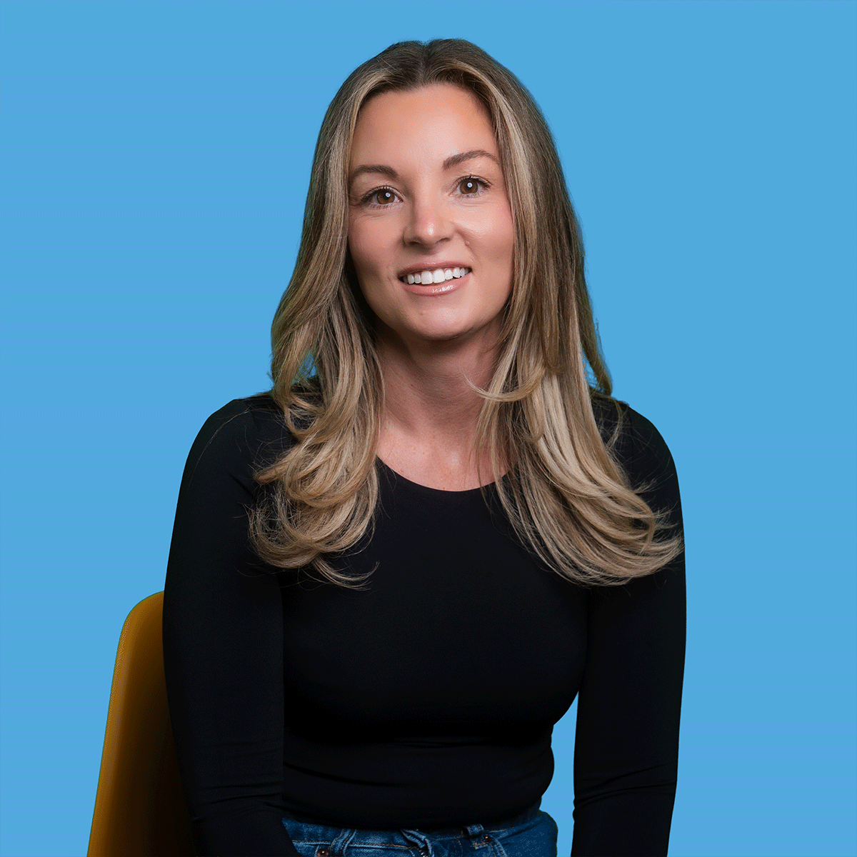 A woman with long blonde hair, wearing a black top, sitting on a yellow chair against a blue background, smiling at the camera.