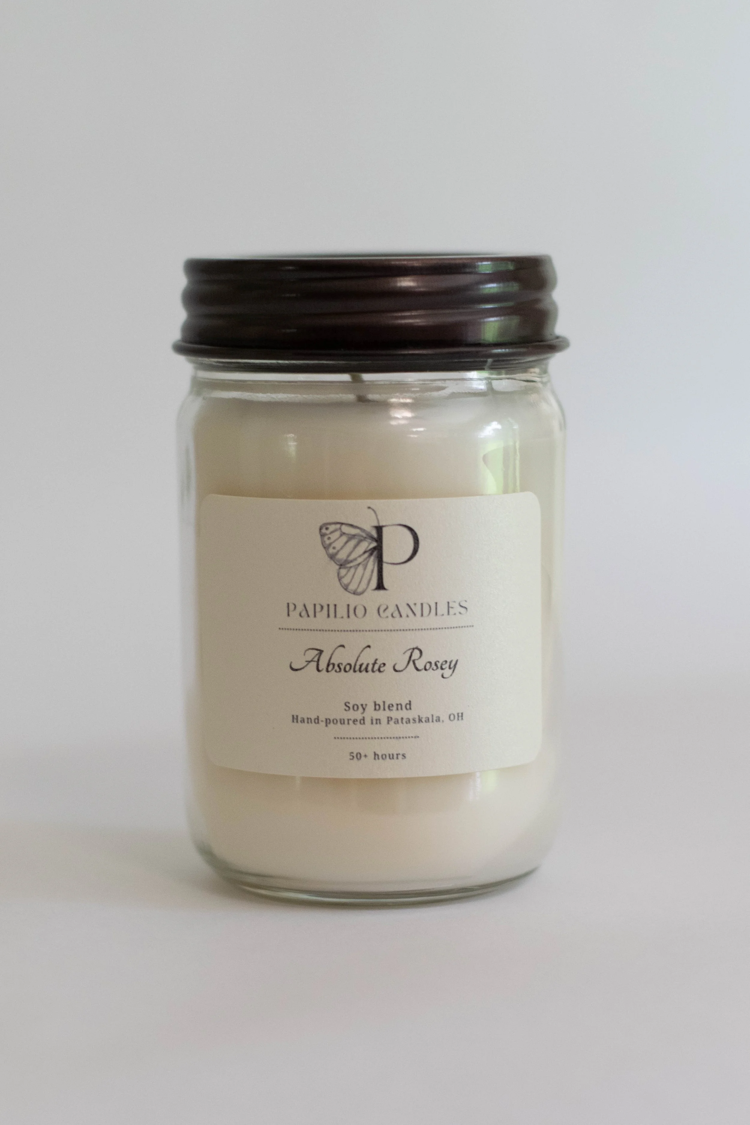 A white candle in a glass jar with a dark metal lid. The label reads 'Papillo Candles Absolute Rosey' and describes it as a soy blend candle hand-poured in Pataskala, Ohio, with an estimated burn time of over 50 hours.