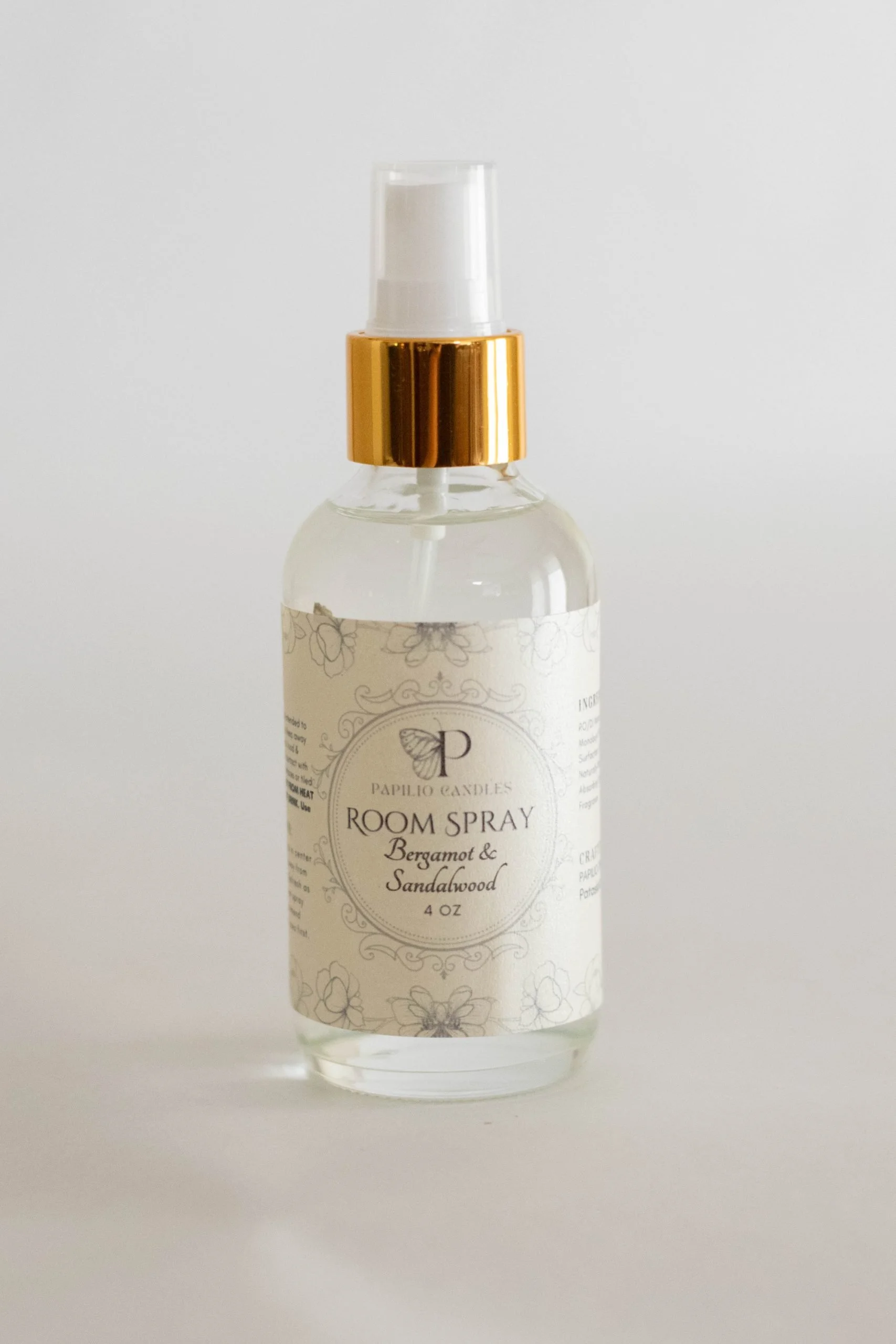 Clear glass room spray bottle with a white label, gold spray nozzle, and a white cap, containing an aromatic product named 'Bergamot & Sandalwood' by Papilio Candles.