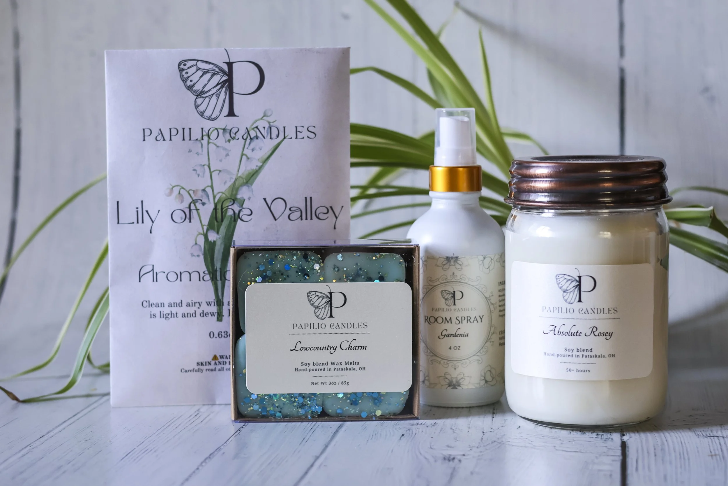 An arrangement of Papilio Candles products with a brochure, soap bars, room spray, and candle jars on a white wooden surface with green leaves in the background.