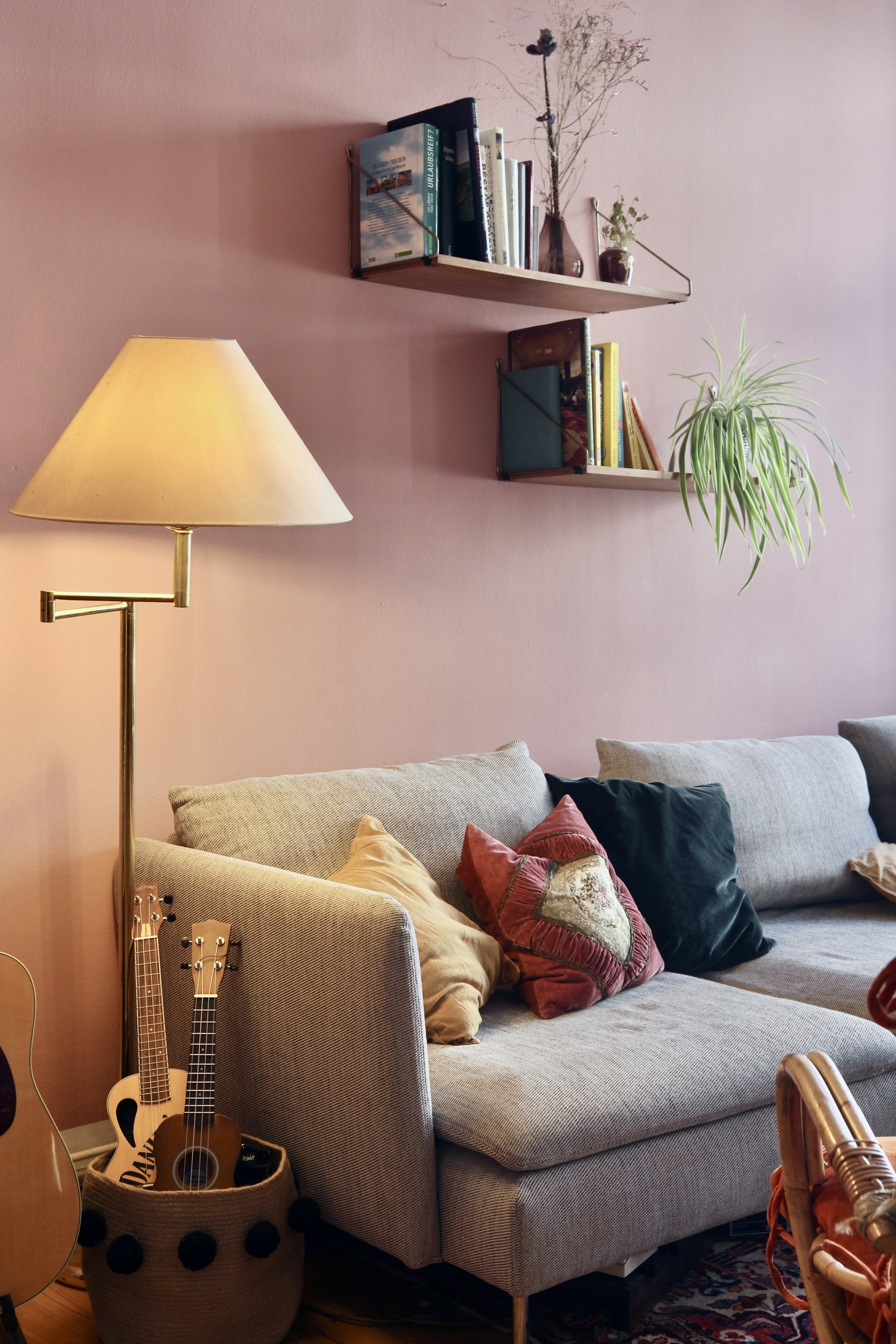 Living room with pink wall, beige sofa with throw pillows, wooden bookshelves with books and plants, standing lamp, and a basket with ukuleles.