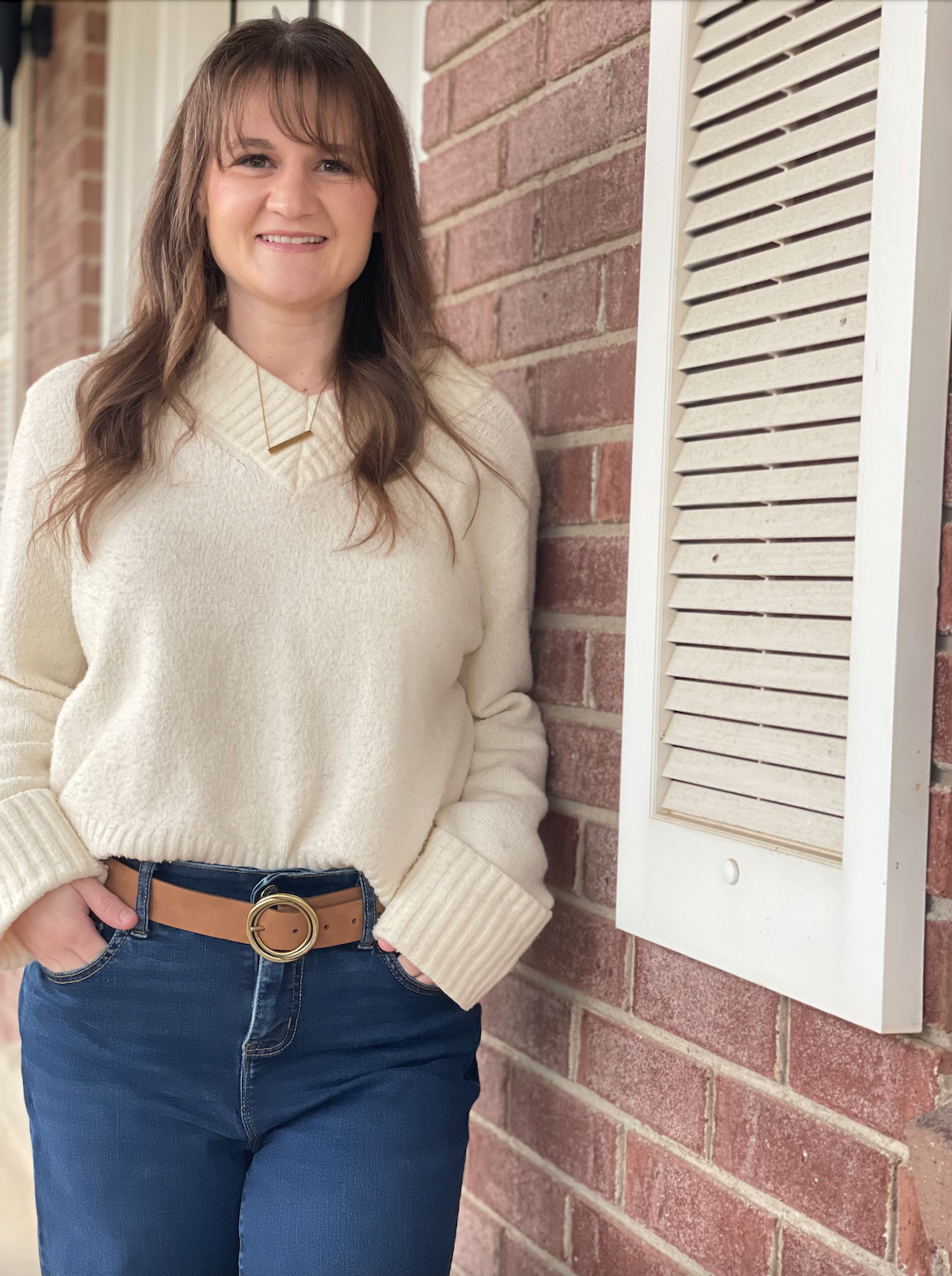 A woman with long brown hair wearing a cream sweater and a tan belt with a gold buckle, standing outside next to a brick wall and a white vent cover, smiling at the camera.