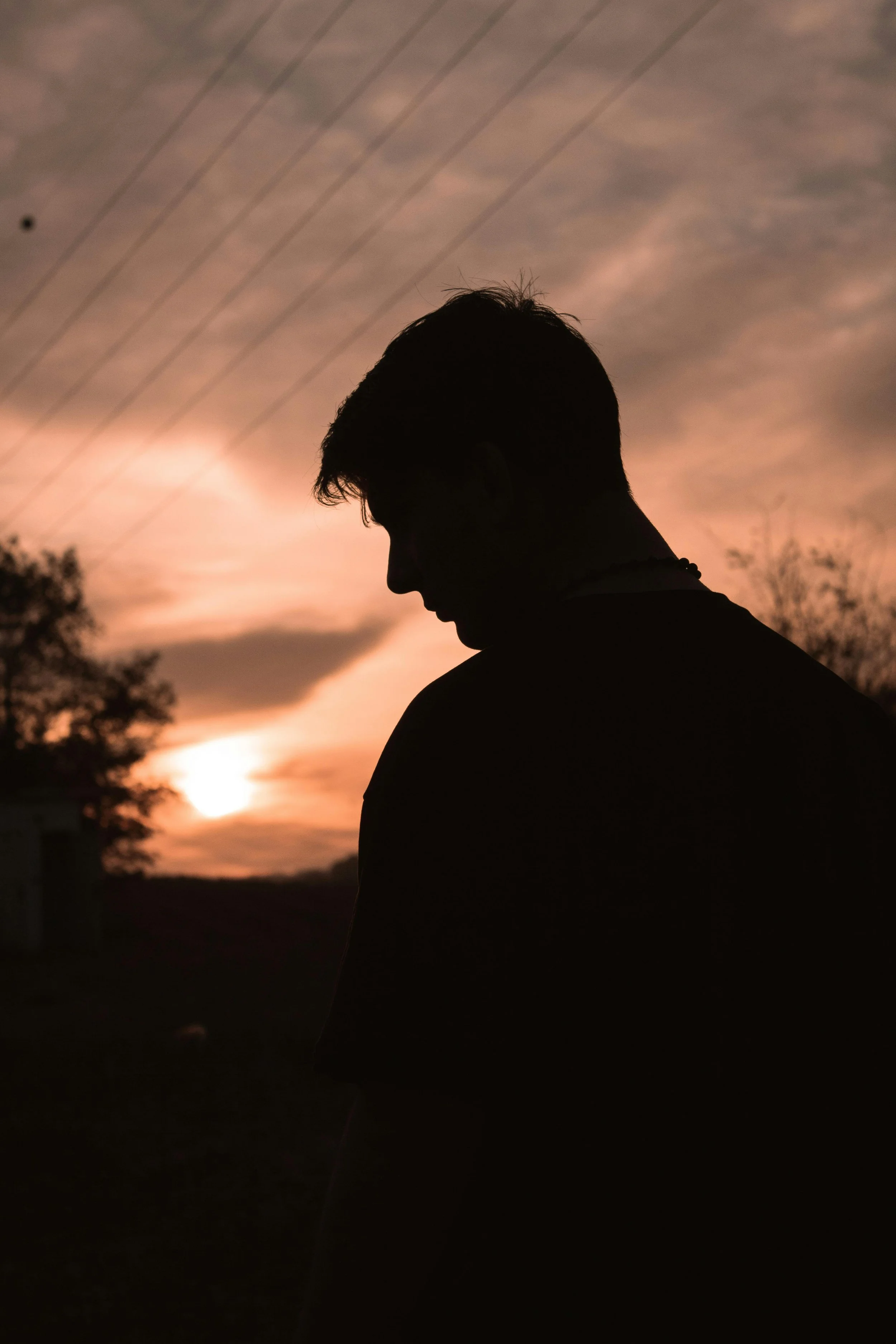 Silhouette of a young man with short hair, looking downward, against a sunset sky with clouds and power lines in the background.