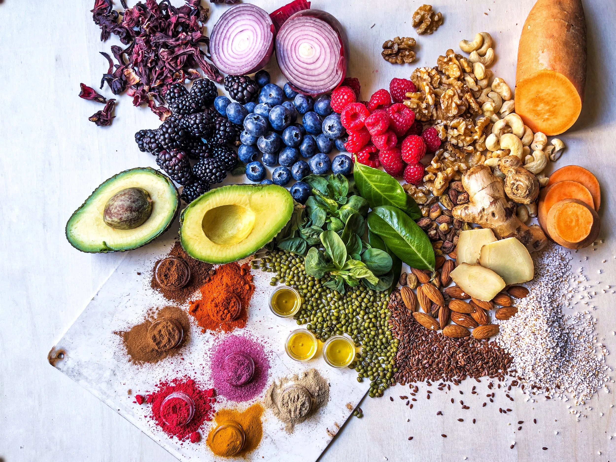 An assortment of fresh fruits, vegetables, nuts, seeds, and spices arranged on a white surface. Includes blueberries, raspberries, blackberries, avocado, red onion, ginger root, sweet potato slices, and various whole and ground spices.