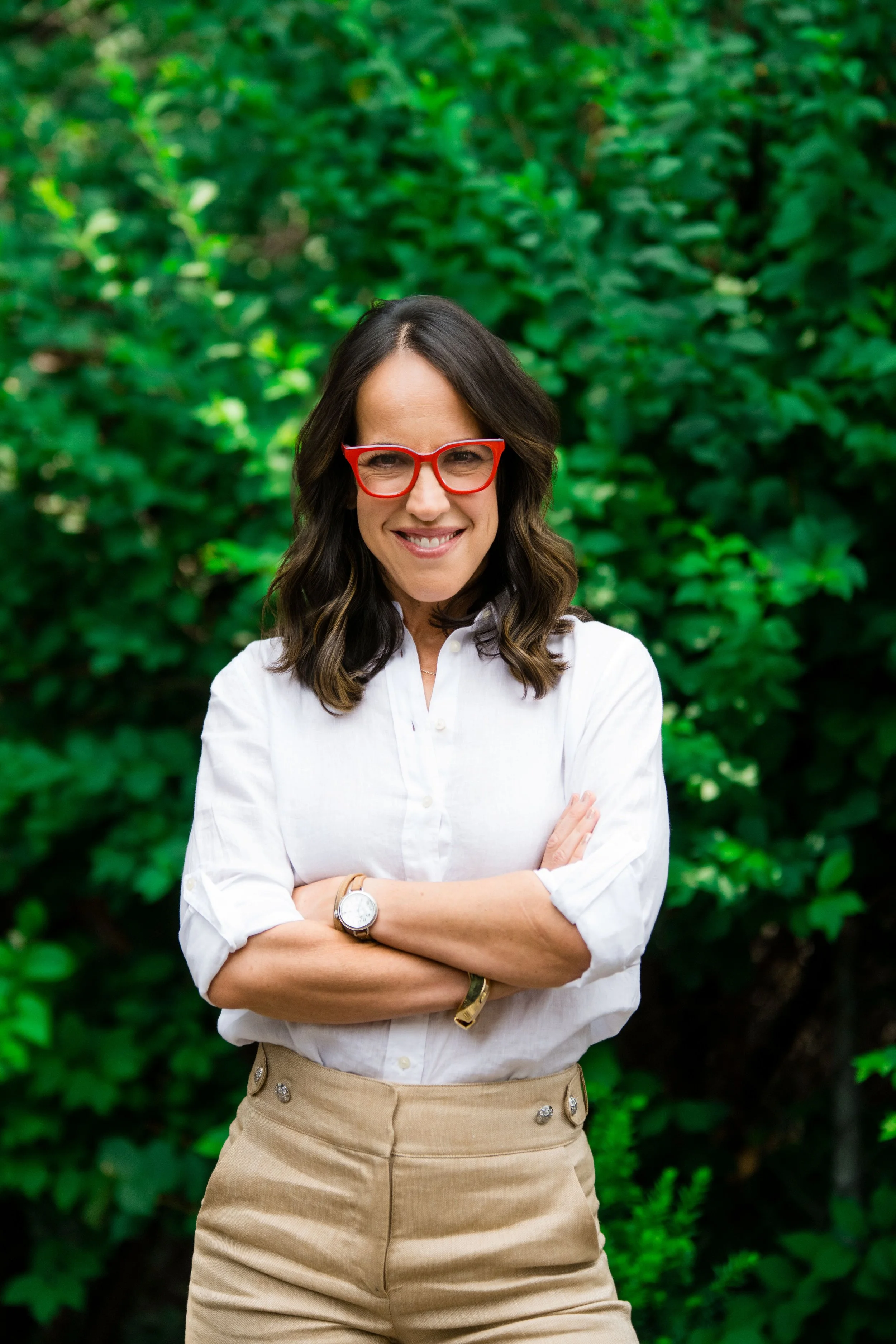 A woman with wavy brown hair, wearing red glasses, a white shirt, khaki pants, and a watch, standing outdoors in front of green foliage, smiling with arms crossed.