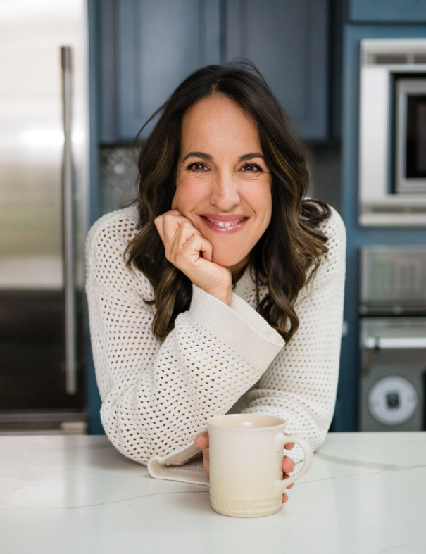 A woman with long dark hair and a white sweater sits at a kitchen counter, smiling and resting her chin on her hand, with a white mug in front of her.