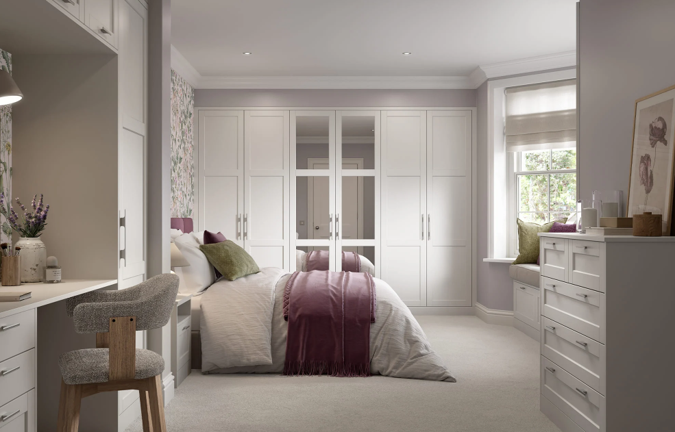 A modern, well-lit bedroom with white built-in wardrobes, a bed with white and purple bedding, window seat with green pillows, and a small white dresser with decorative items.