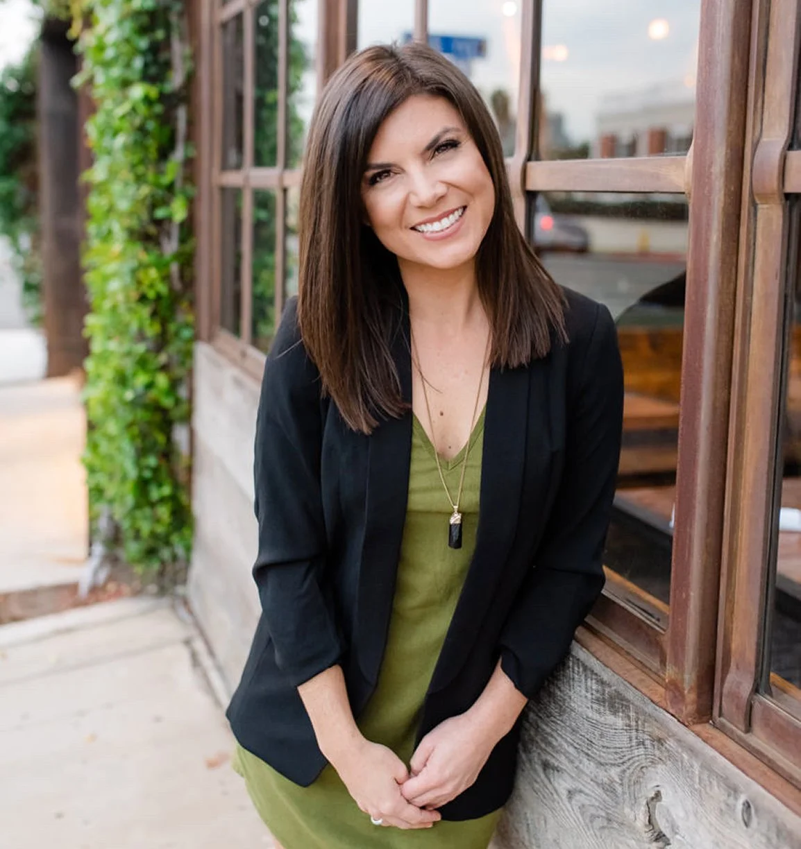 A smiling woman with shoulder-length dark brown hair, wearing a black blazer over an olive green dress, standing outdoors next to a wooden and glass window, with greenery and buildings in the background.