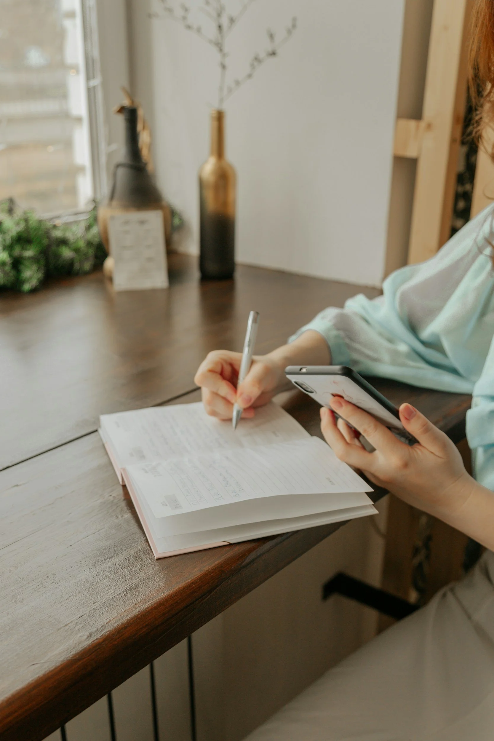 Person taking notes with a pen and smartphone on a wooden table, with a notebook open, behind a window, decorative vases, and a plant in the background.