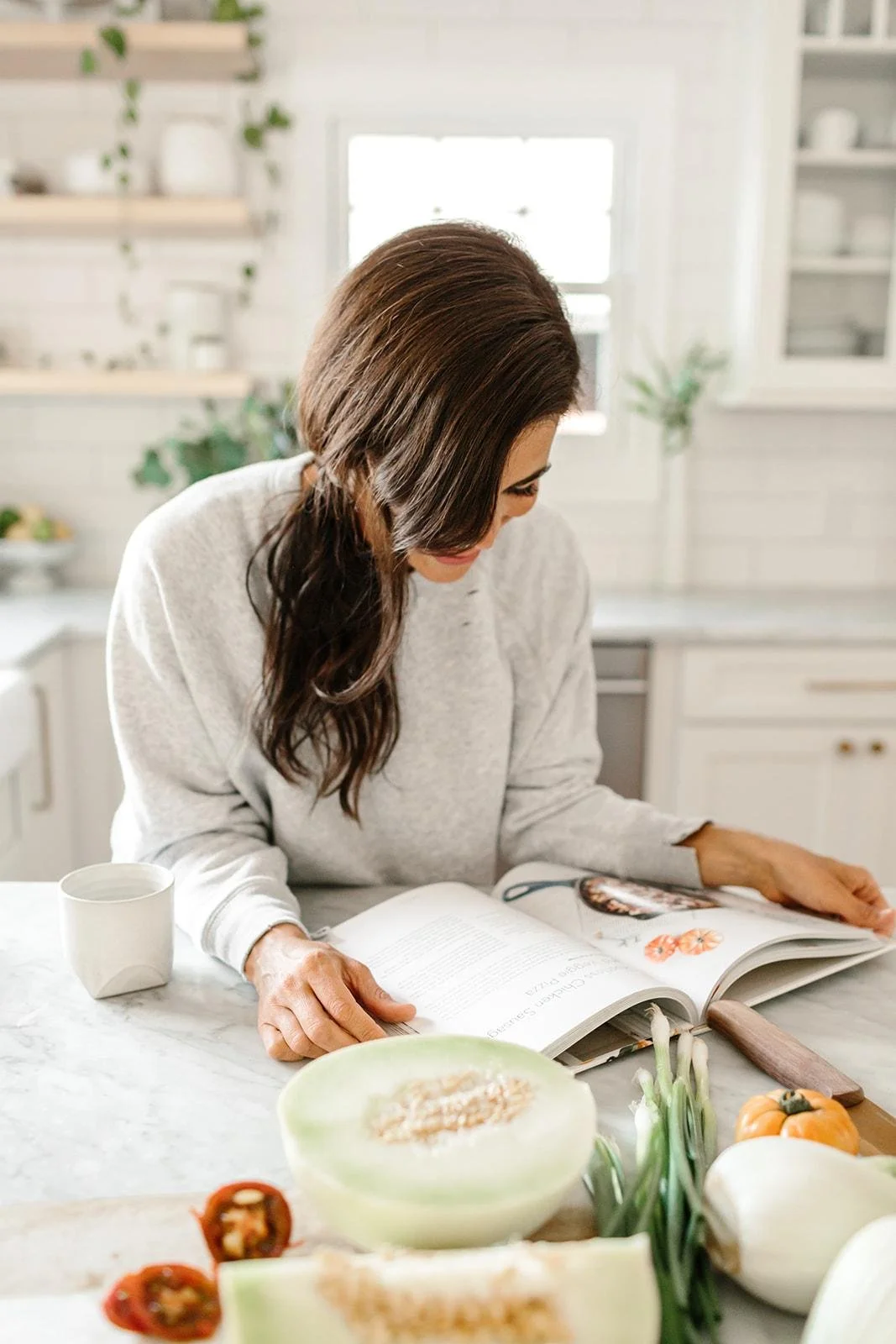 A woman with long brown hair reading a cookbook at a kitchen counter surrounded by vegetables and a mug.