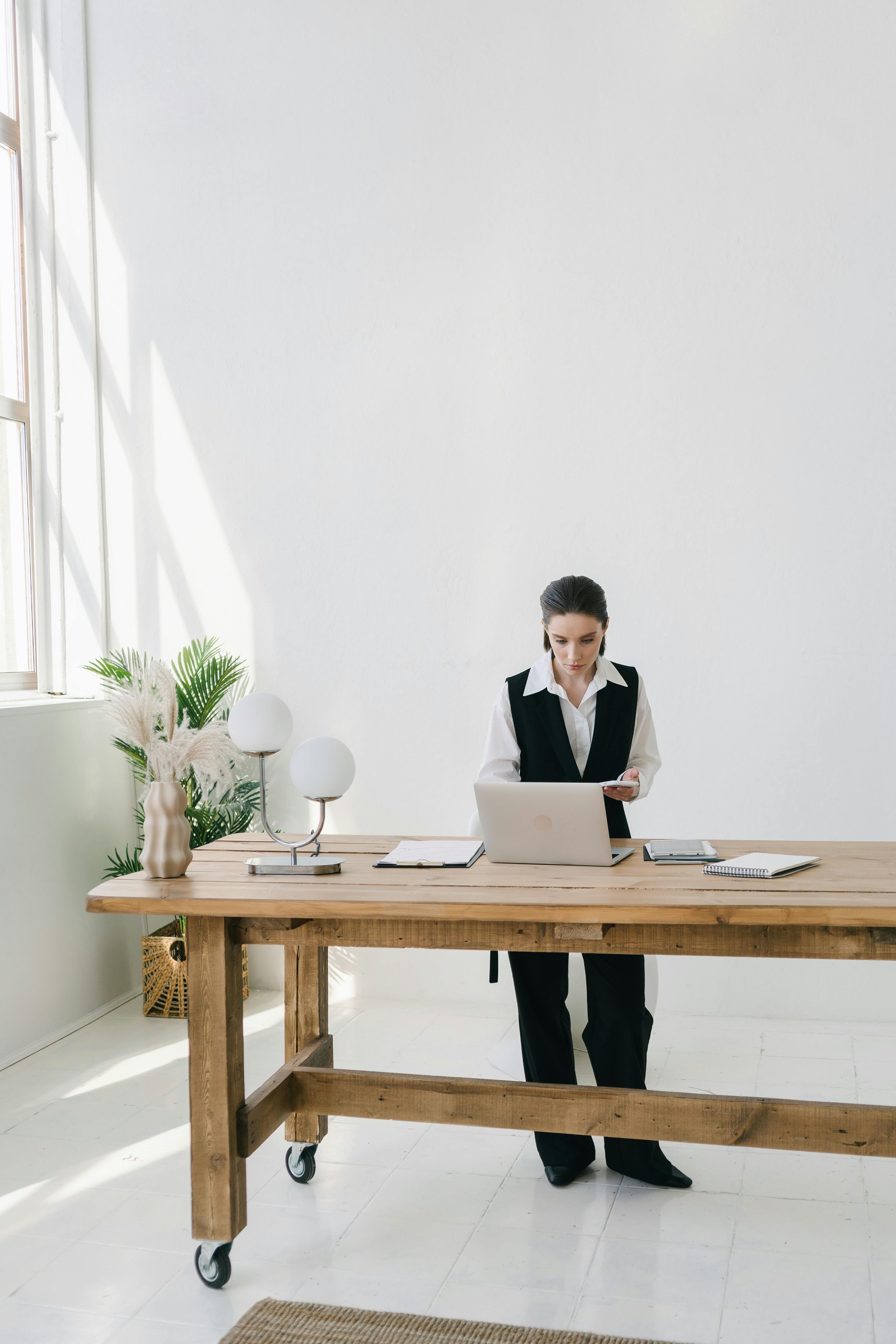 A woman in business attire standing at a wooden desk in a bright, minimalist office, looking at her smartphone and with a laptop, notebooks, and a plant on the desk.