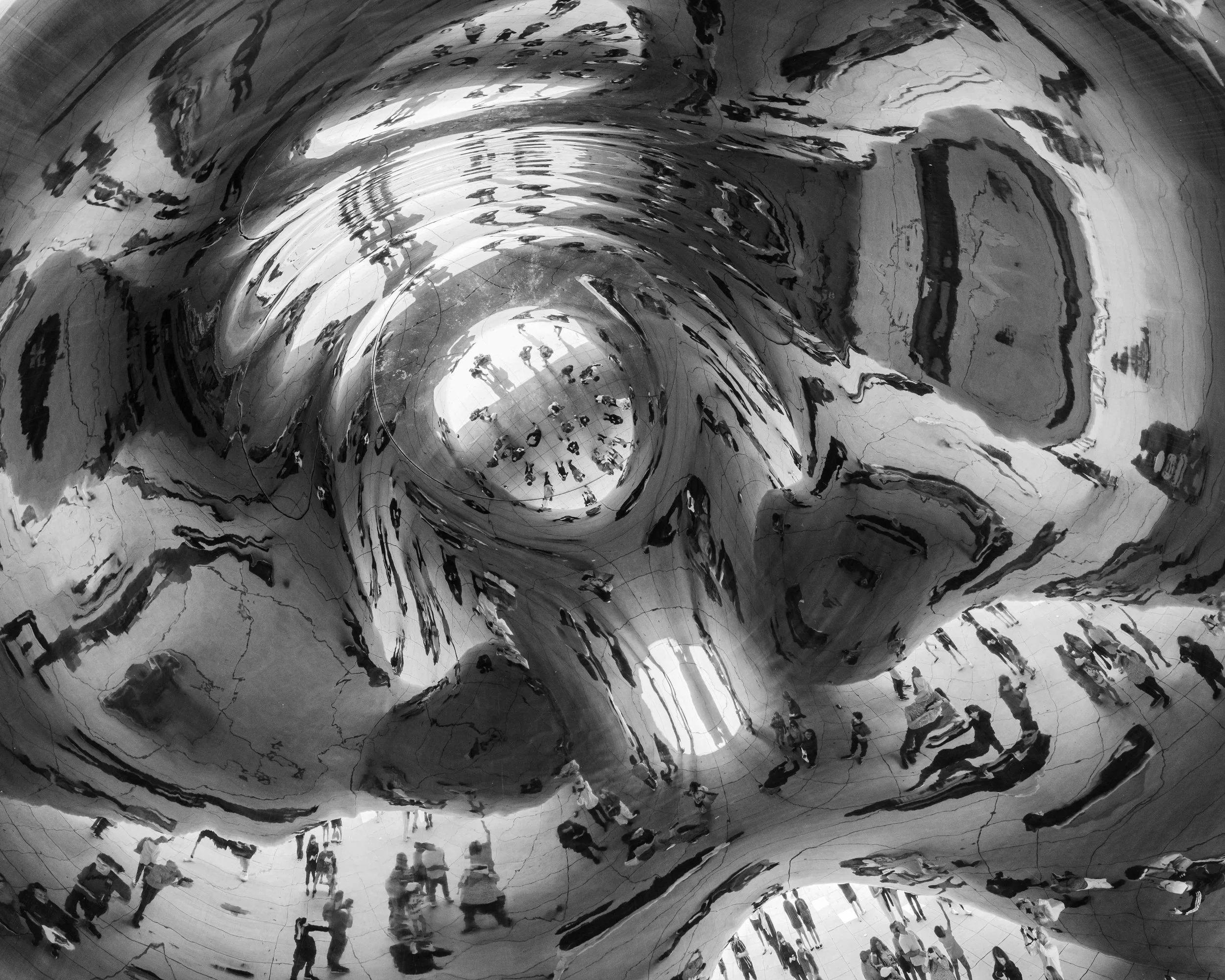 Reflections of people walking in the interior of a shiny, metallic, curved structure, seen from below.