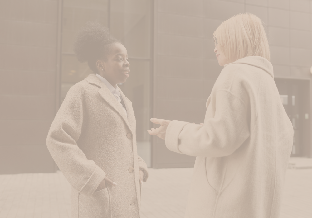 Two women having a conversation indoors, both wearing beige coats.