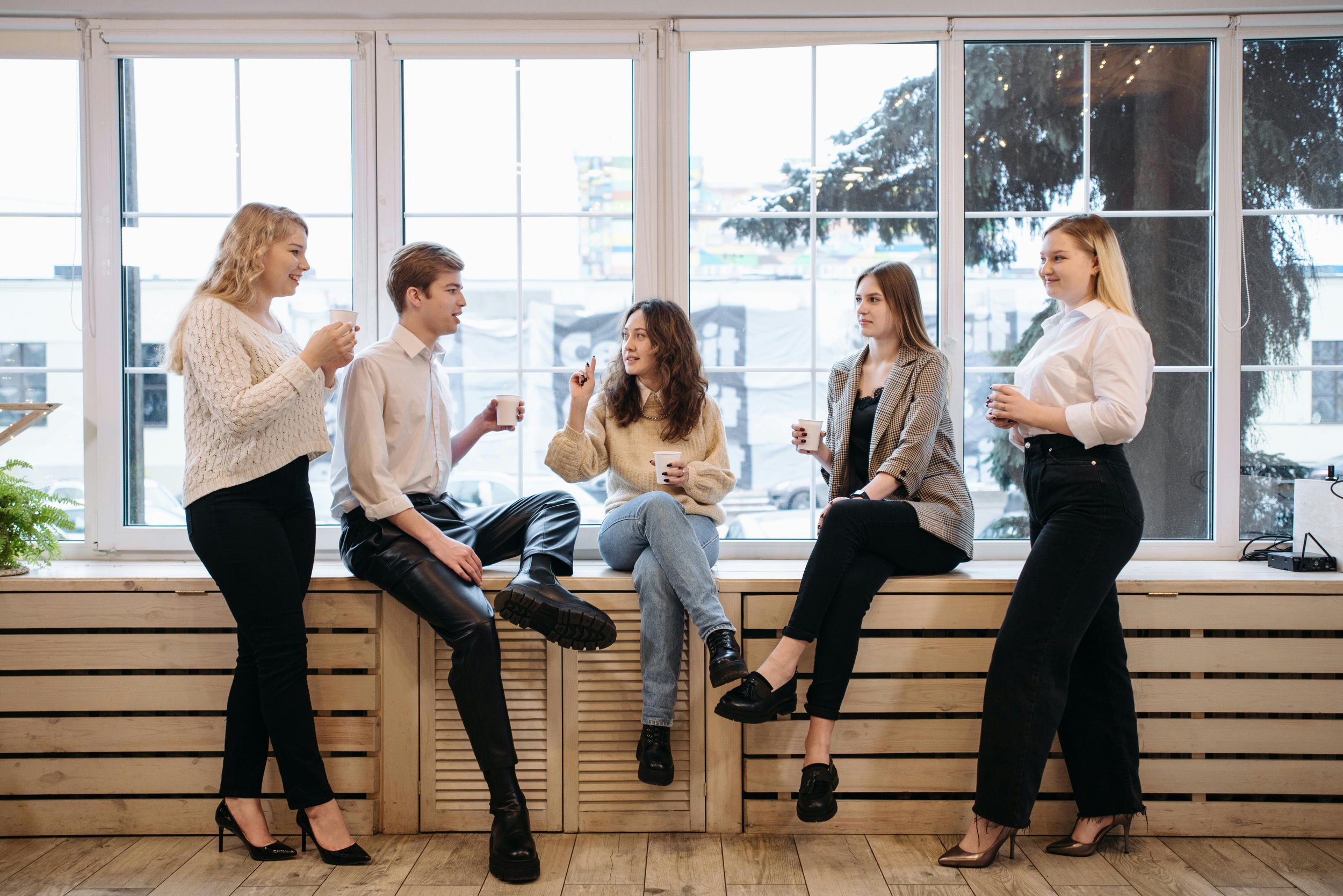 Five young women and men having a conversation while sitting and standing by a large window with a city view, holding cups.
