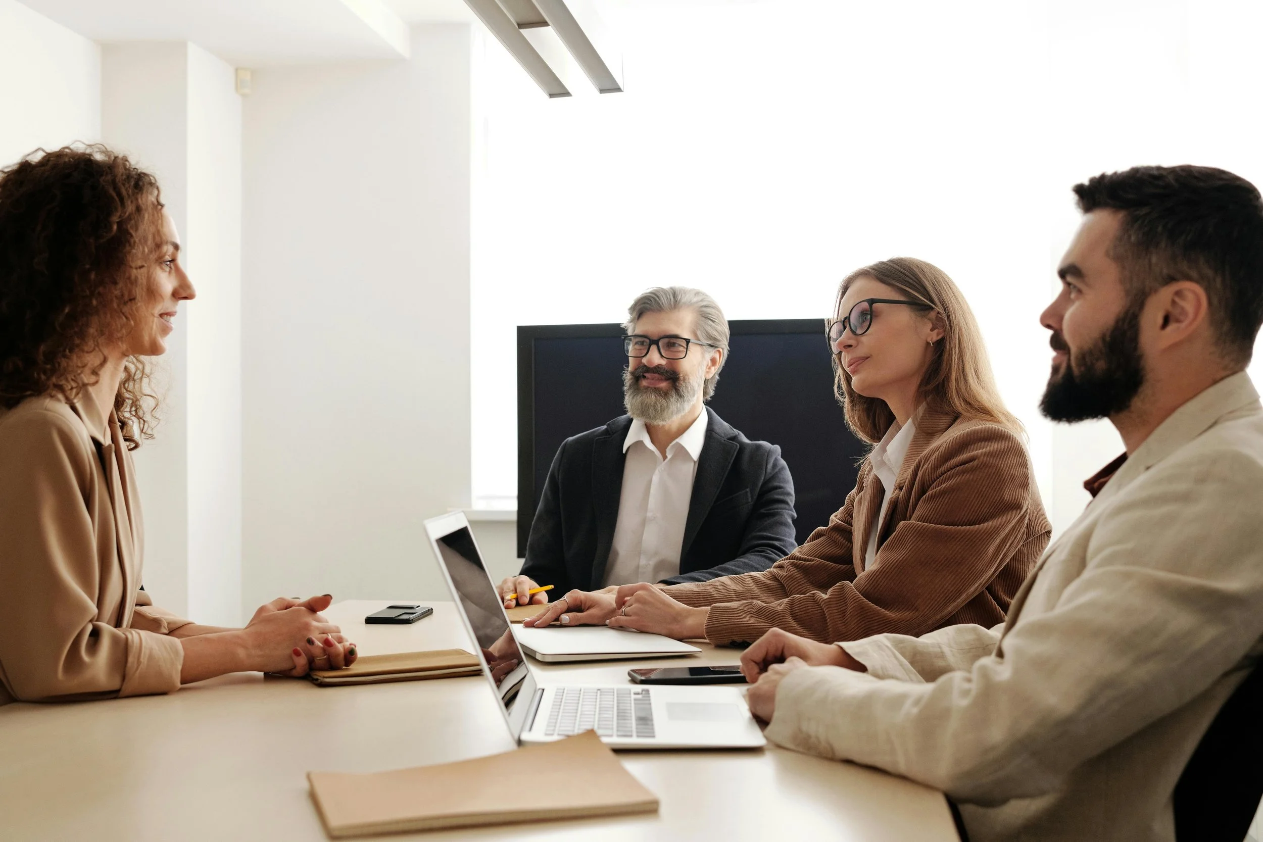 Four business professionals, two men and two women, sitting at a conference table during a meeting, with laptops and notebooks in front of them, in a bright office environment.