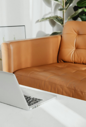A white laptop on a white table in front of a tan leather sofa in a modern living room with plants.