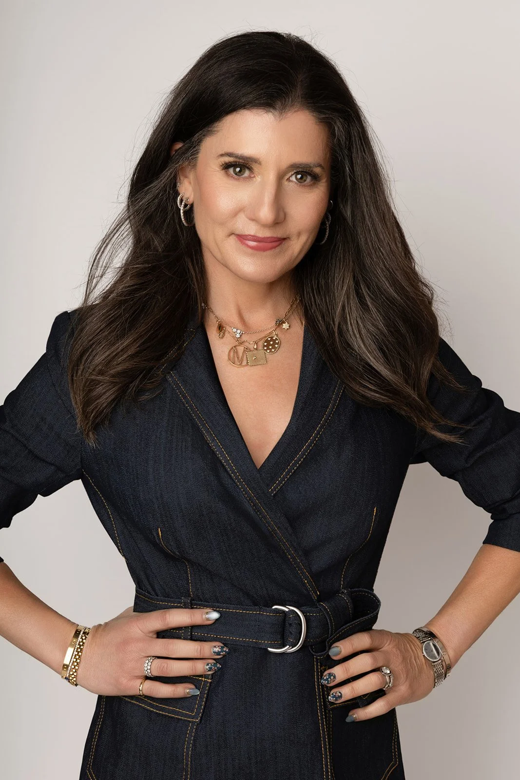A woman with brown hair, wearing a dark denim dress and gold jewelry, posing with her hands on her hips against a neutral background.