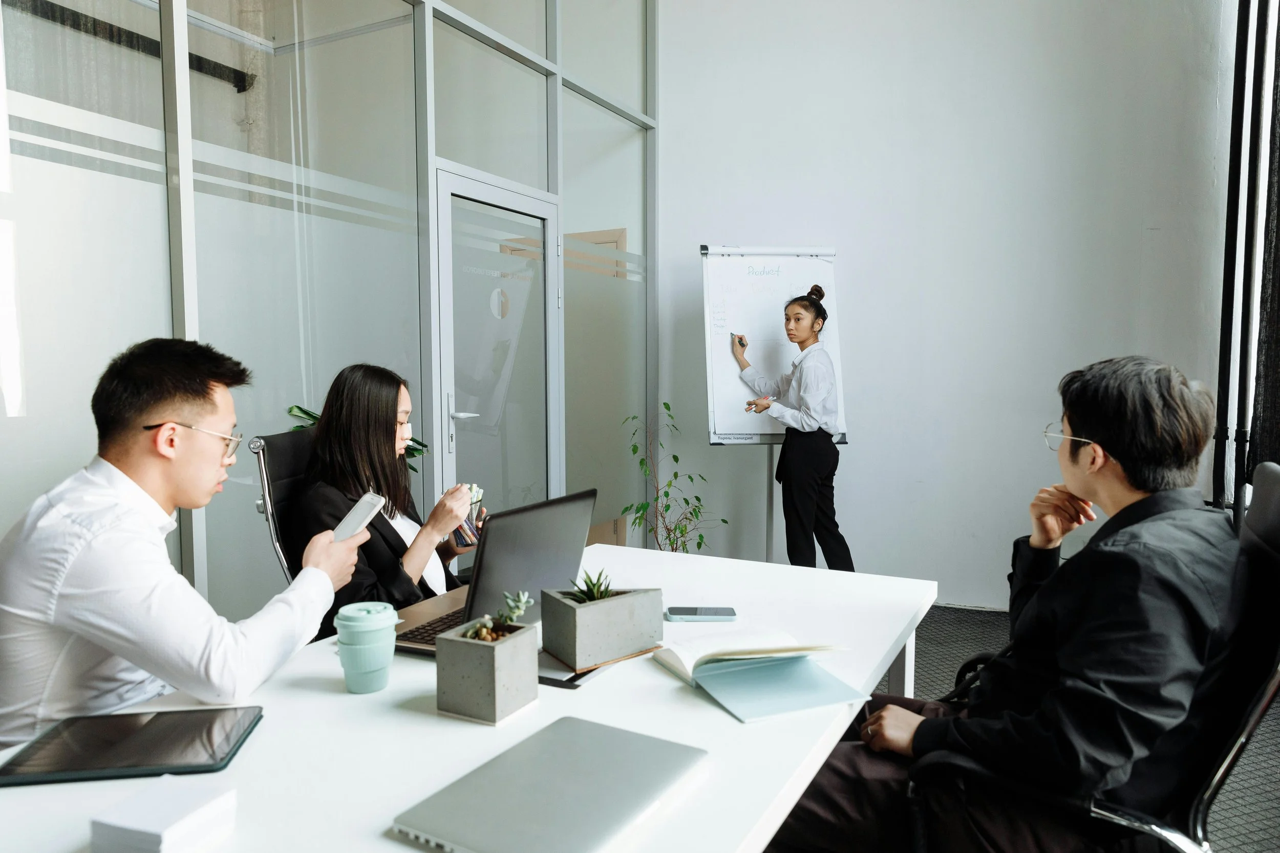 Business meeting with four people in a modern conference room, one woman is standing at a flip chart, three people seated at the table examining documents or electronic devices.