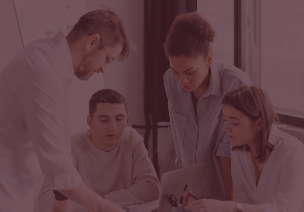 Four people collaborating on a laptop in an office or classroom setting.