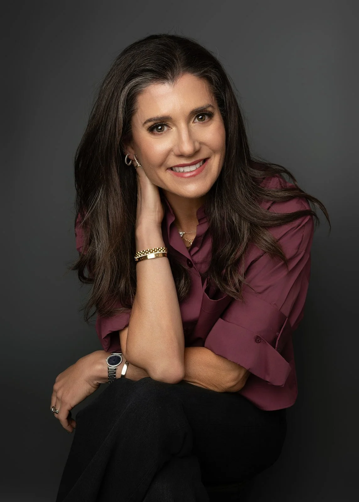 A woman with long brown hair wearing a purple blouse, smiling, sitting against a dark gray background.