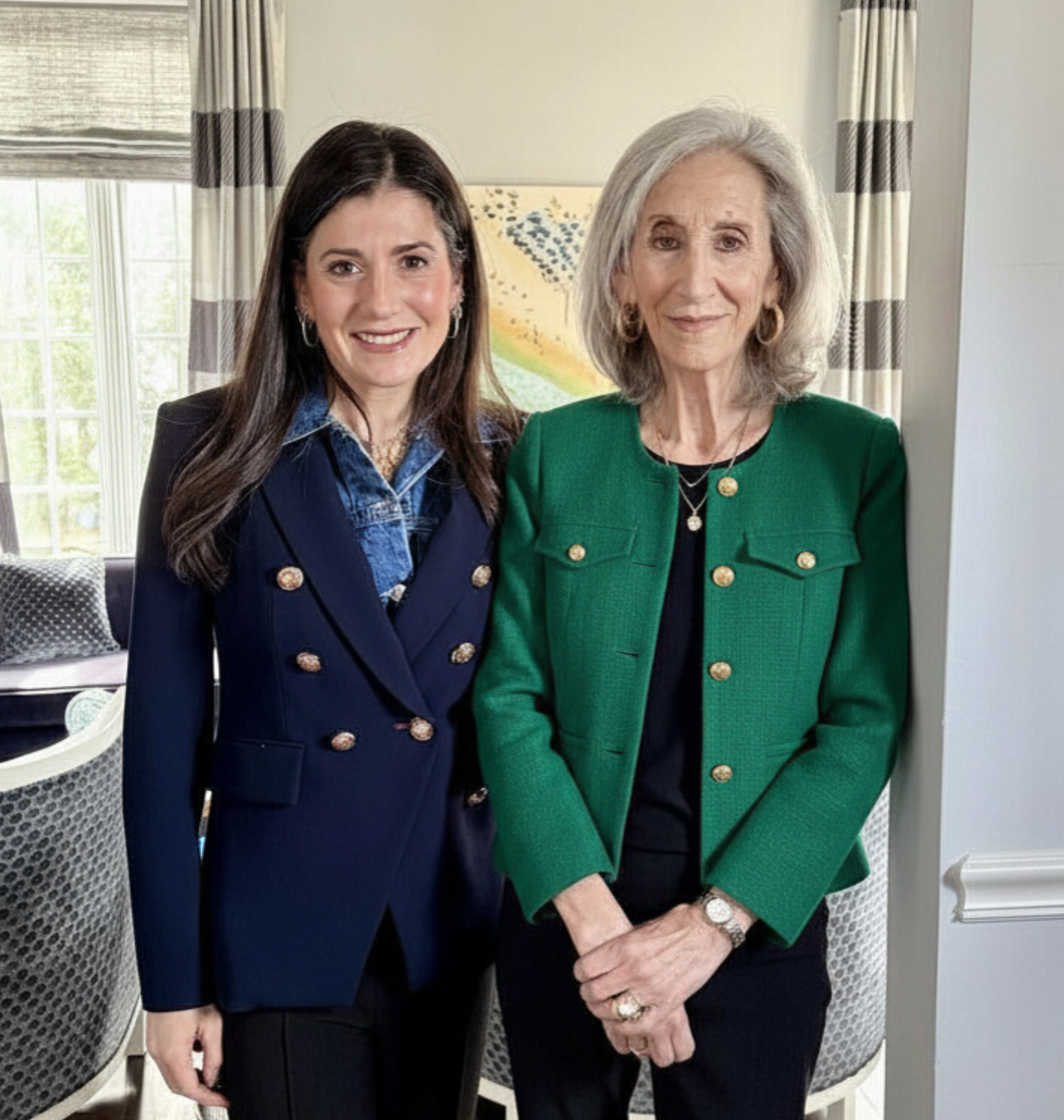 Two women standing together indoors, smiling at the camera. The woman on the left has dark hair, wearing a navy blue blazer with gold buttons, and a denim shirt underneath. The woman on the right has gray hair, wearing a green blazer with gold buttons, a black top, and accessorized with gold jewelry.