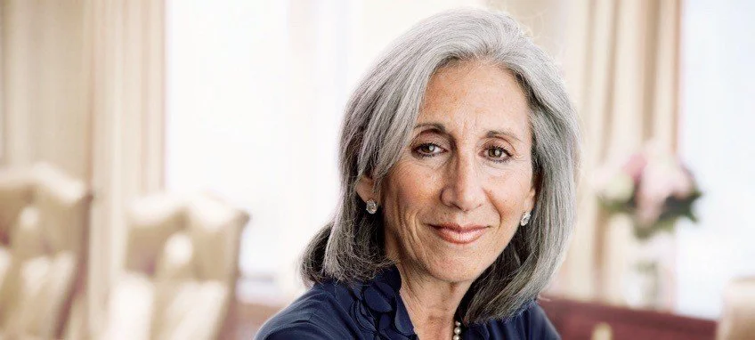 Portrait of a senior woman with gray hair, wearing a navy blue top and pearl earrings, smiling softly in a well-lit room with beige curtains and floral decorations in the background.