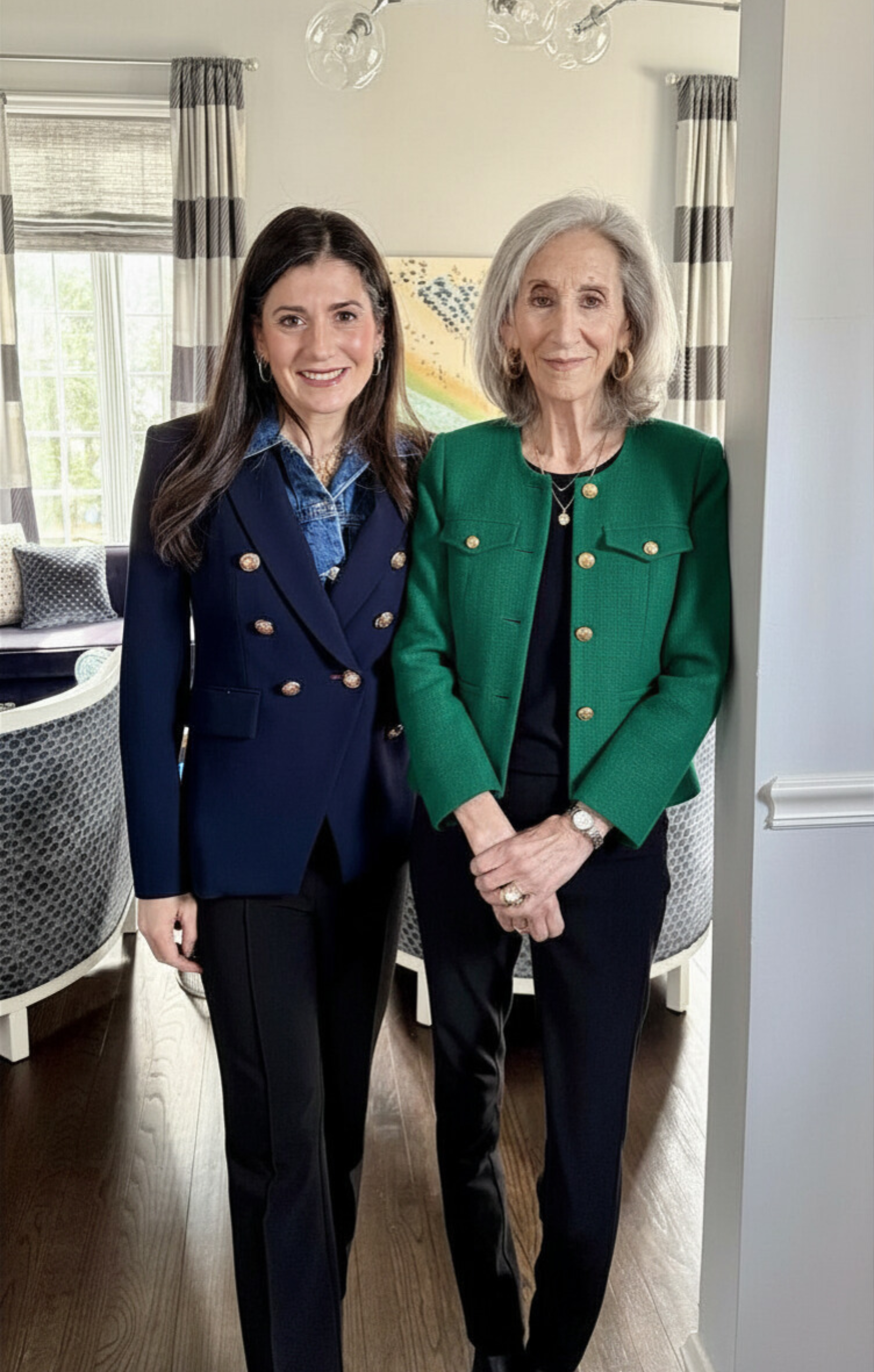 Two women standing side by side in a well-lit room with large windows, curtains, and artwork in the background. The woman on the left has long dark hair and is wearing a navy blazer with gold buttons, a denim shirt, and black pants. The woman on the right has shoulder-length gray hair and is dressed in a green blazer with gold buttons, a black top, and black pants. Both women are smiling.
