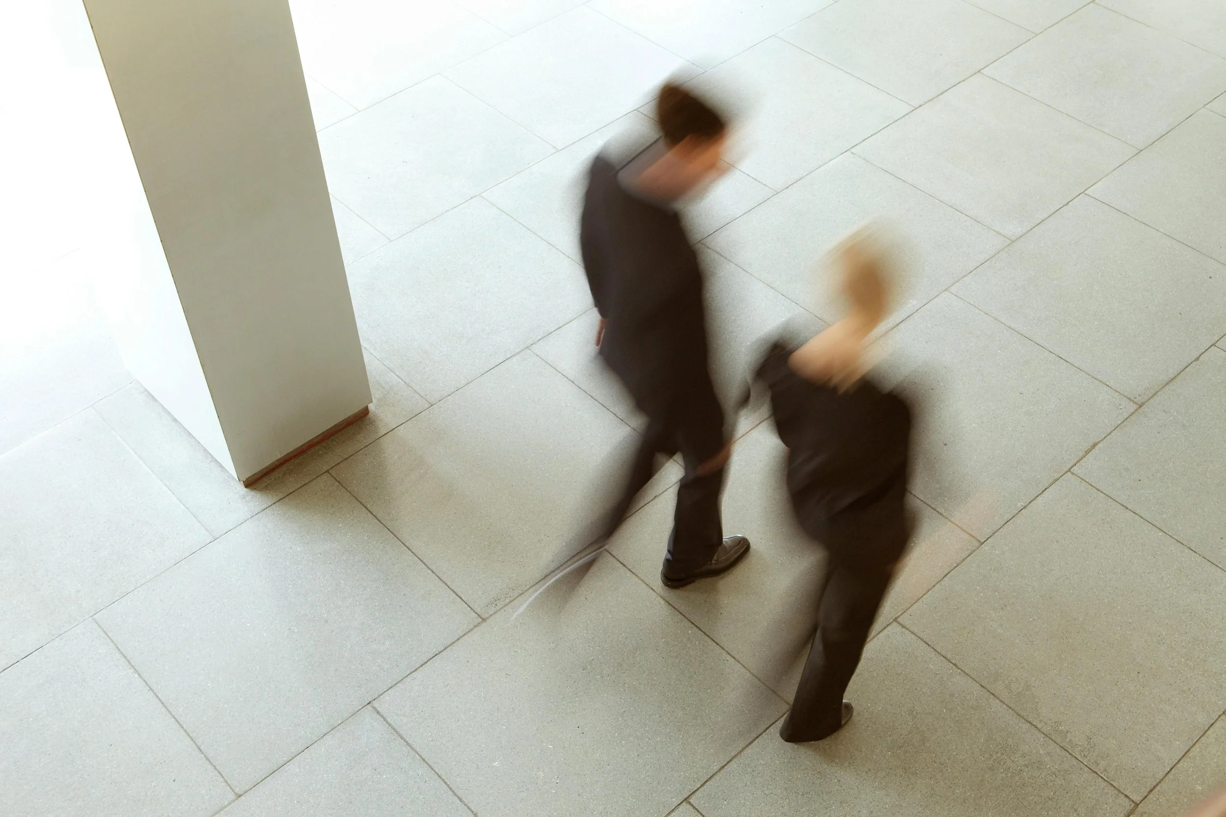 Two people in business attire standing and bending over on a tiled floor in an indoor setting, with a white wall or pillar nearby and motion blur indicating movement.