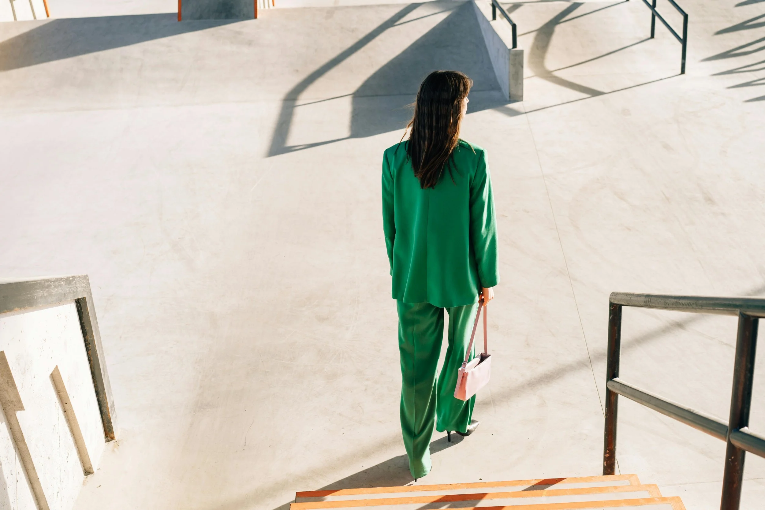 A woman dressed in a bright green suit walking down stairs in a modern, minimalistic outdoor space, carrying a pink handbag.