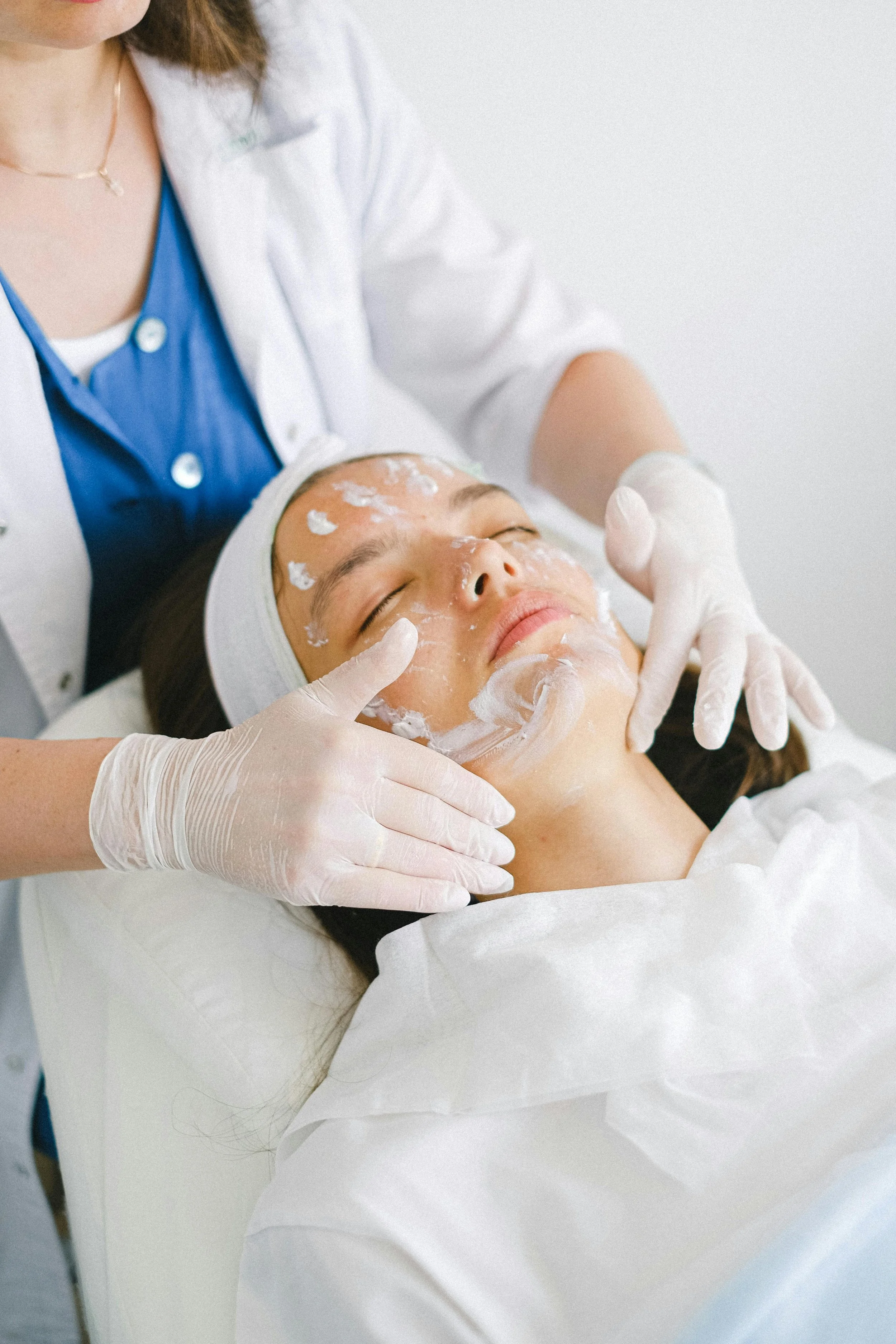 A woman is receiving a facial treatment from a professional wearing white gloves at a spa or clinic. The woman is lying on a treatment bed with her eyes closed, and a facial mask is being applied to her face.