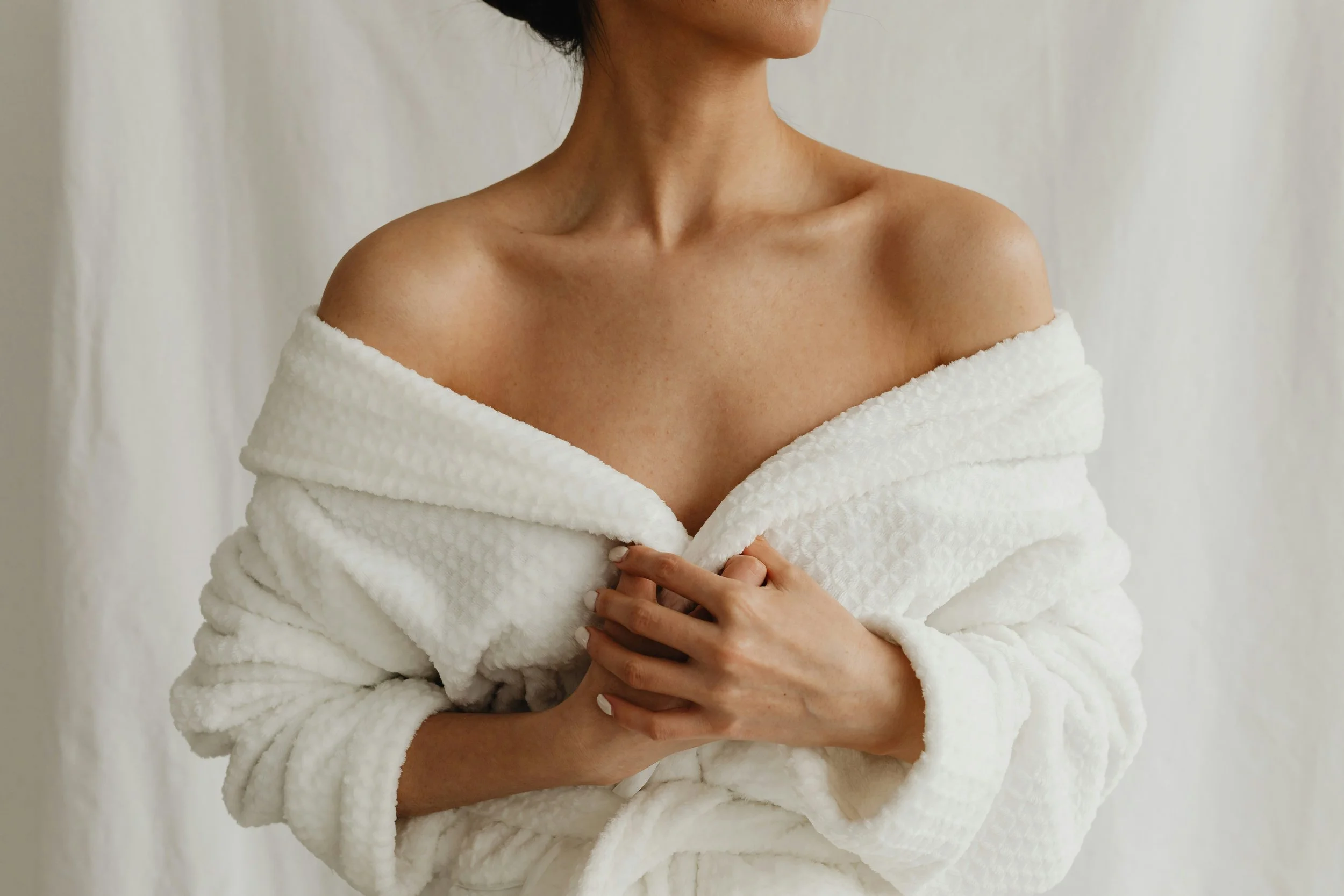 A woman wearing a white textured bathrobe, holding it closed near her chest, with bare shoulders and neck visible, standing against a light background.