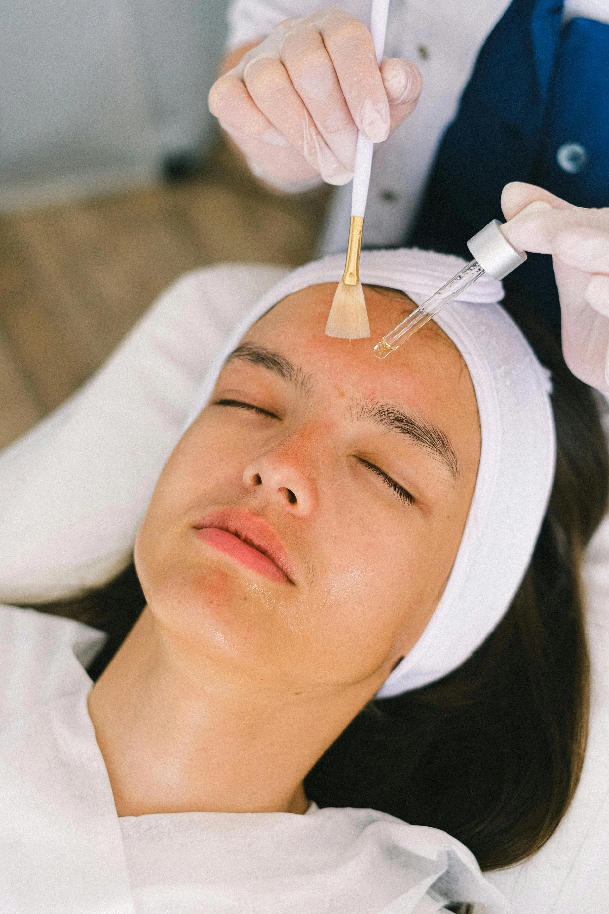 A woman with closed eyes receiving a facial treatment, with a skincare professional applying serum with a dropper and using a brush on her forehead.