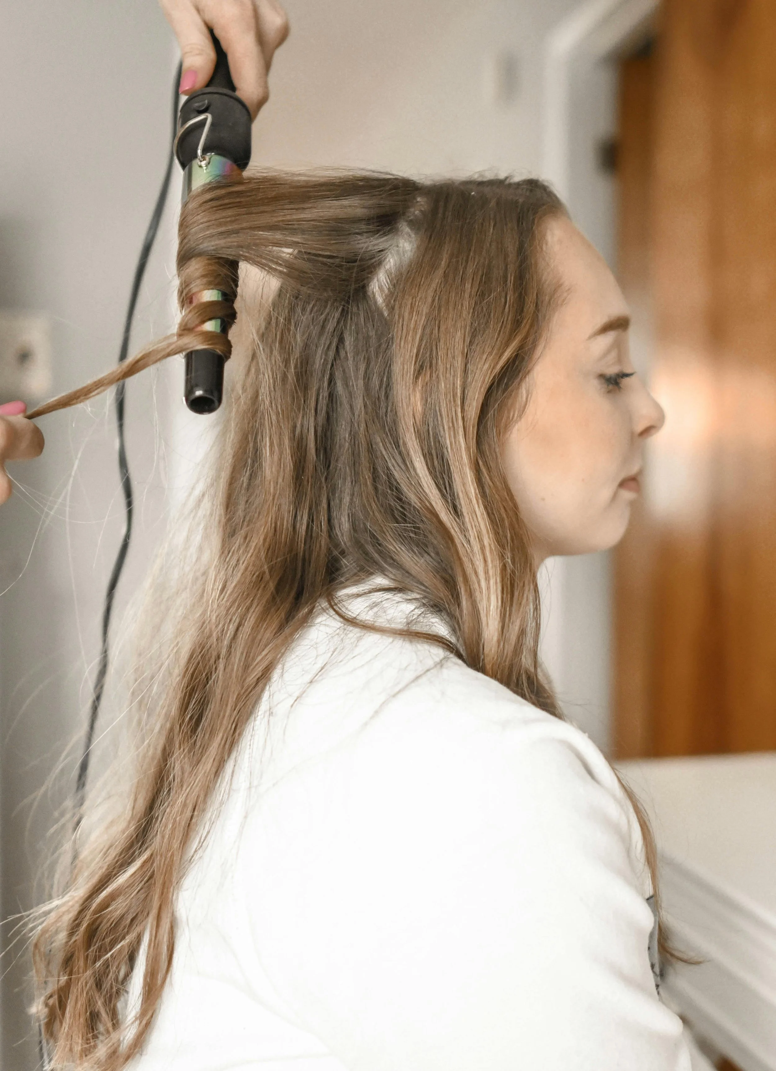 A woman with long brown hair having her hair curled with a curling iron by a hairstylist.