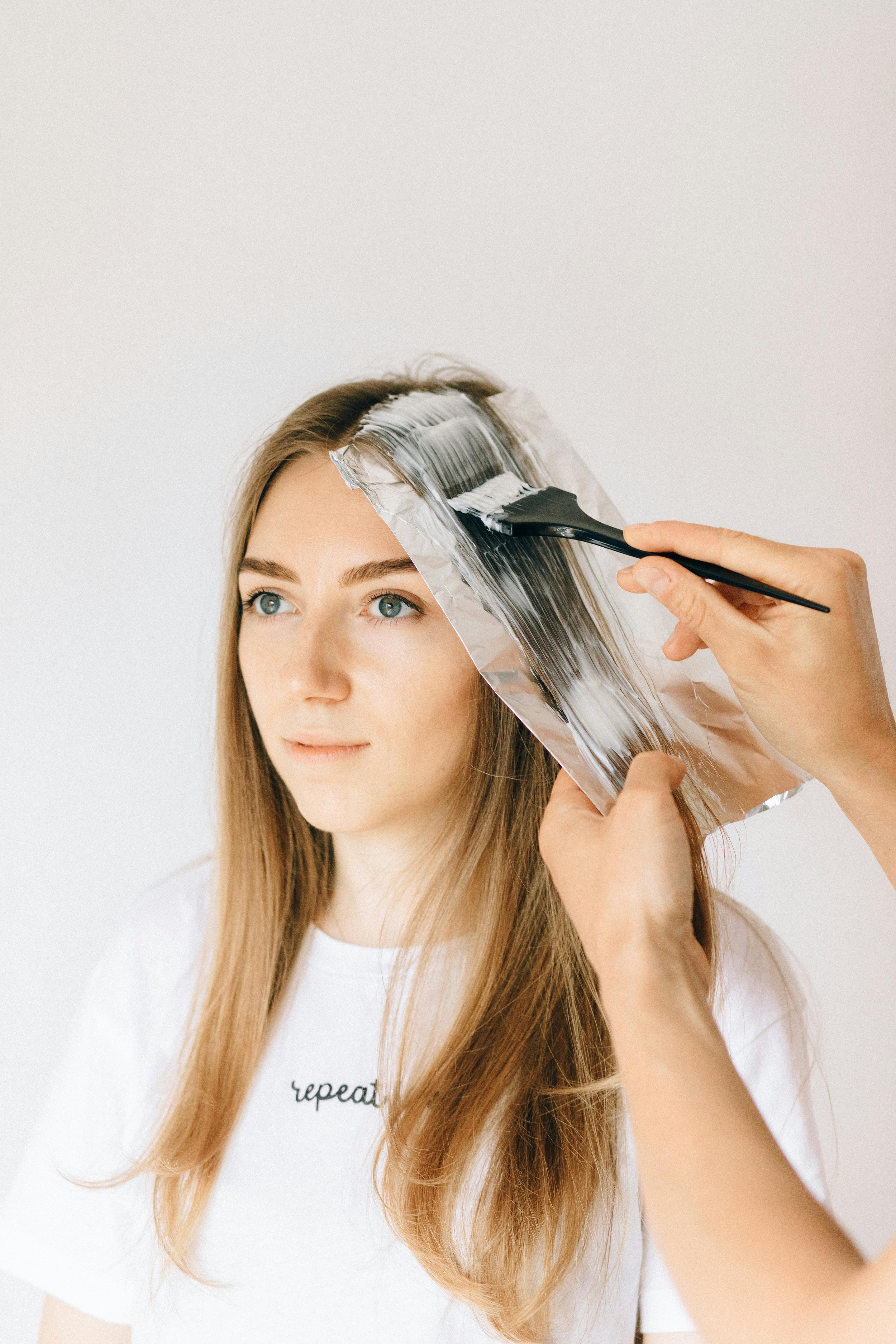 A woman with long, light brown hair gets hair highlights applied using foils and a brush by a stylist.