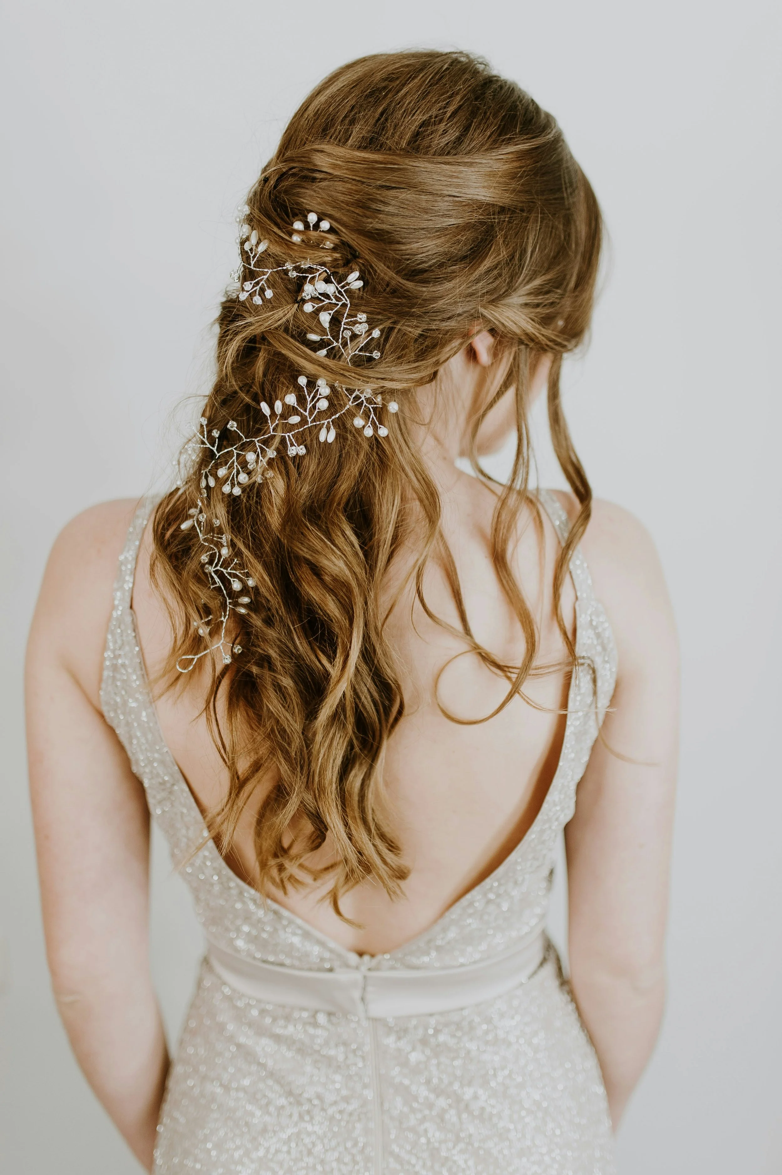 A woman with long, wavy brown hair decorated with a white floral hairpiece, wearing a sparkly sleeveless gown with an open back.