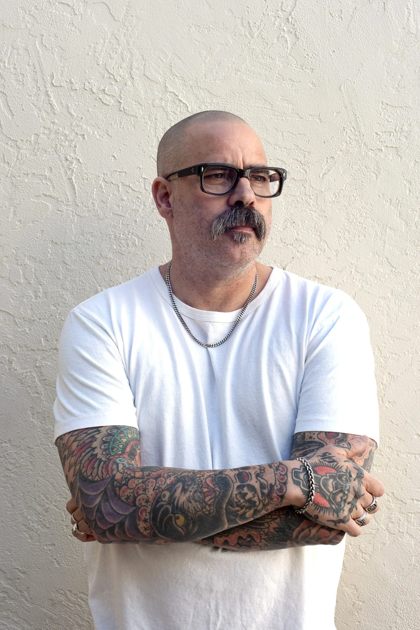 Man with glasses, bald head, and a mustache and beard, wearing a white t-shirt, silver chain, and multiple rings, standing with arms crossed against a textured beige wall.