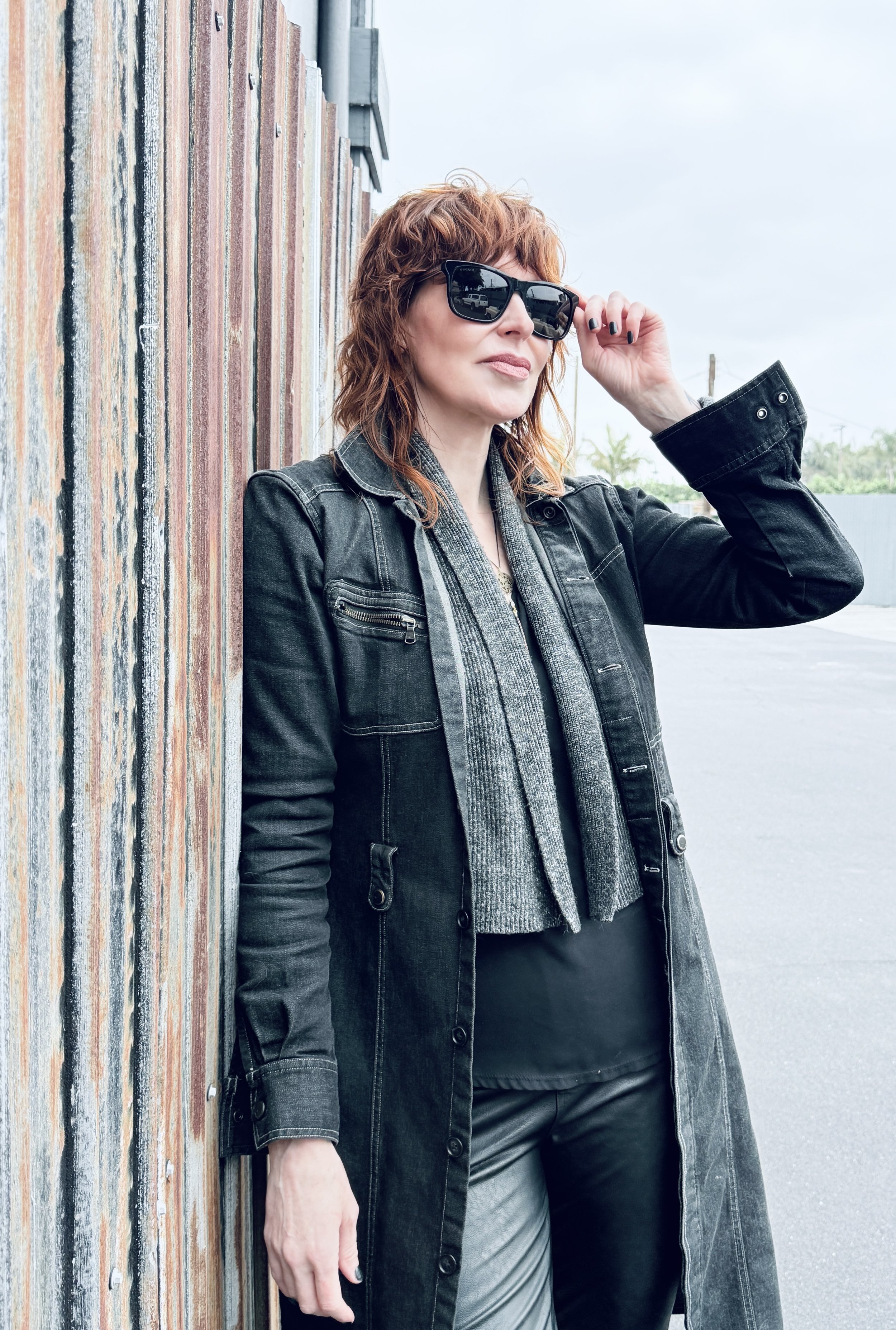 A woman with red hair and sunglasses leaning against a rusty metal wall outdoors on a cloudy day, wearing a black denim coat, a gray sweater, and dark pants.