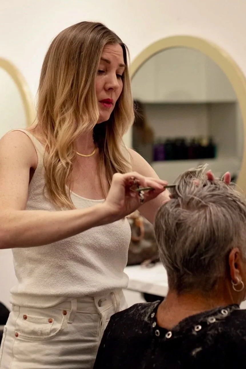 A woman with long, wavy blonde hair getting her hair styled by a stylist in a salon. The stylist is holding a section of the woman's hair with a pair of scissors or styling tool.