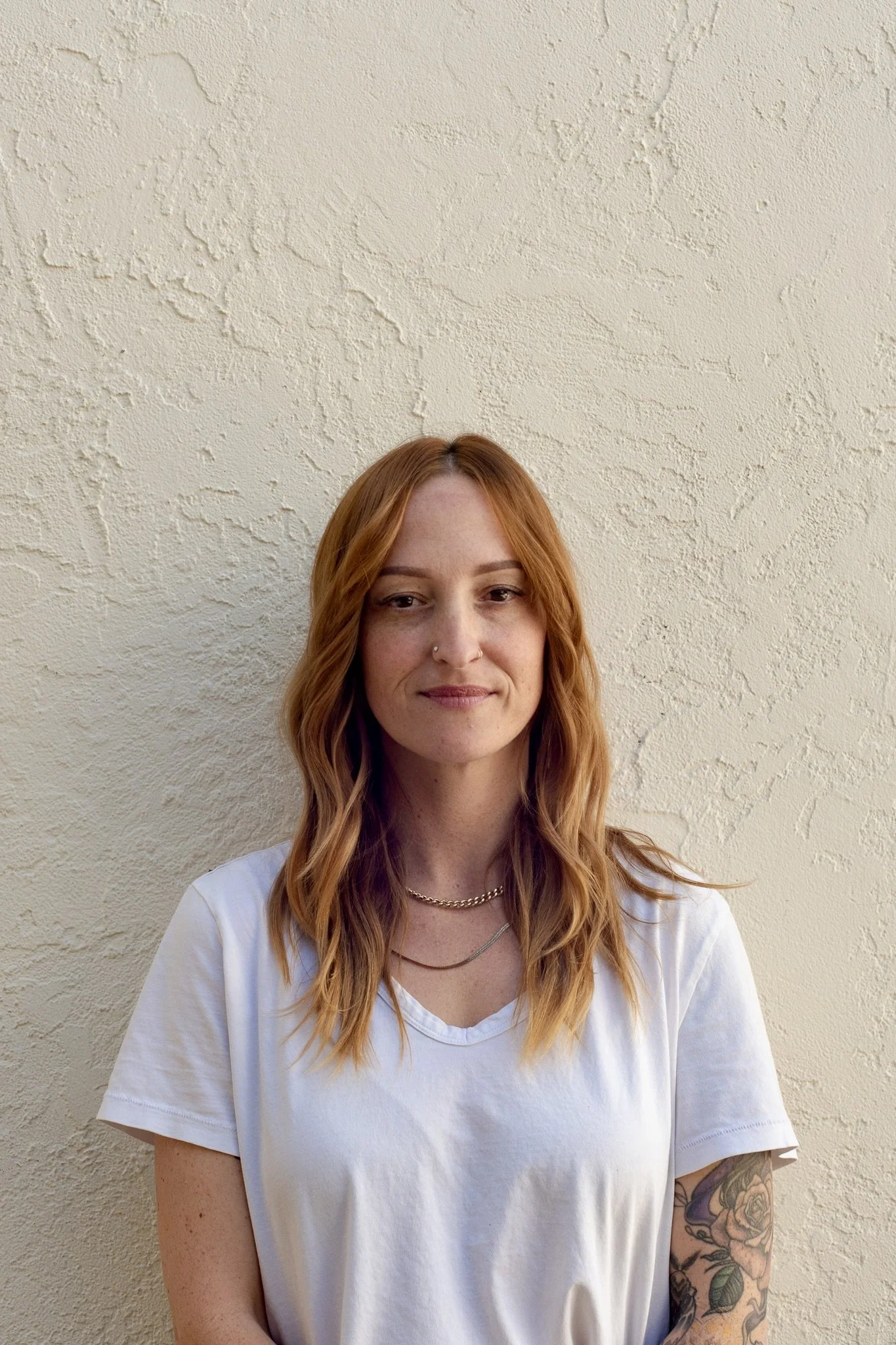 A woman with long, wavy red hair and a nose piercing, wearing a white T-shirt and layered necklaces, standing against a textured beige wall.