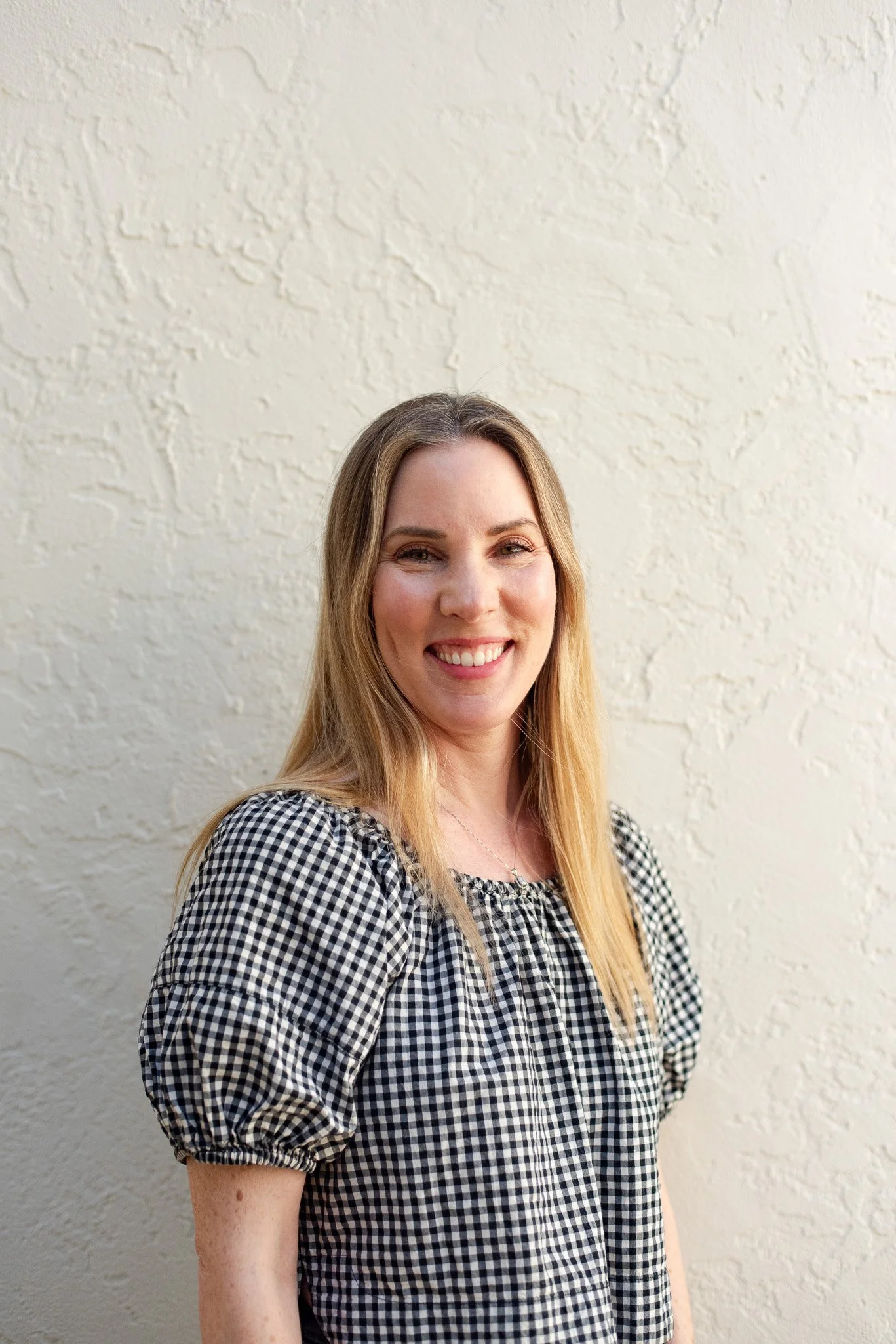 Woman with blonde hair smiling, wearing a black and white checkered blouse, standing against a textured white wall.