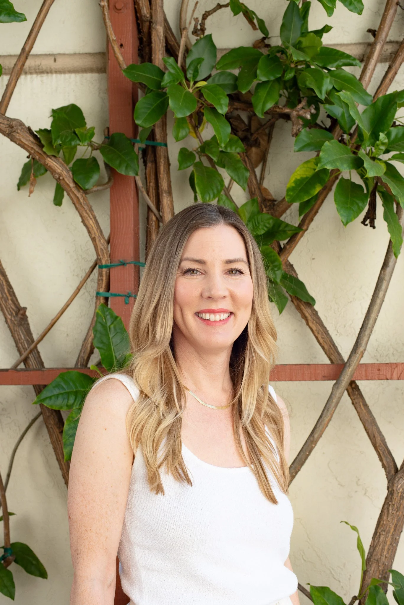 A woman with long blonde hair, smiling, wearing a white sleeveless top and a gold necklace, standing outdoors in front of a wall with green leafy plants and brown wooden trellises.