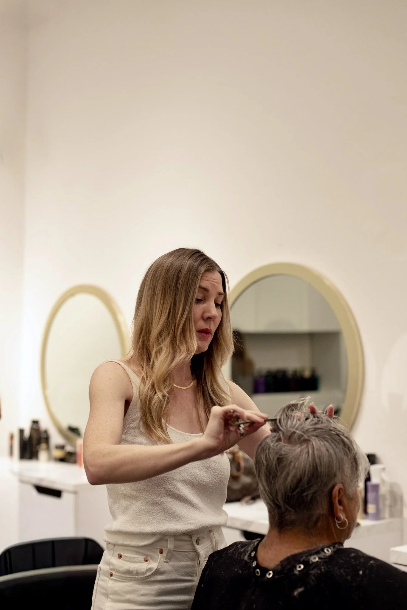 A woman with blonde hair is cutting the hair of another woman who has short gray hair, in a salon or barbershop.