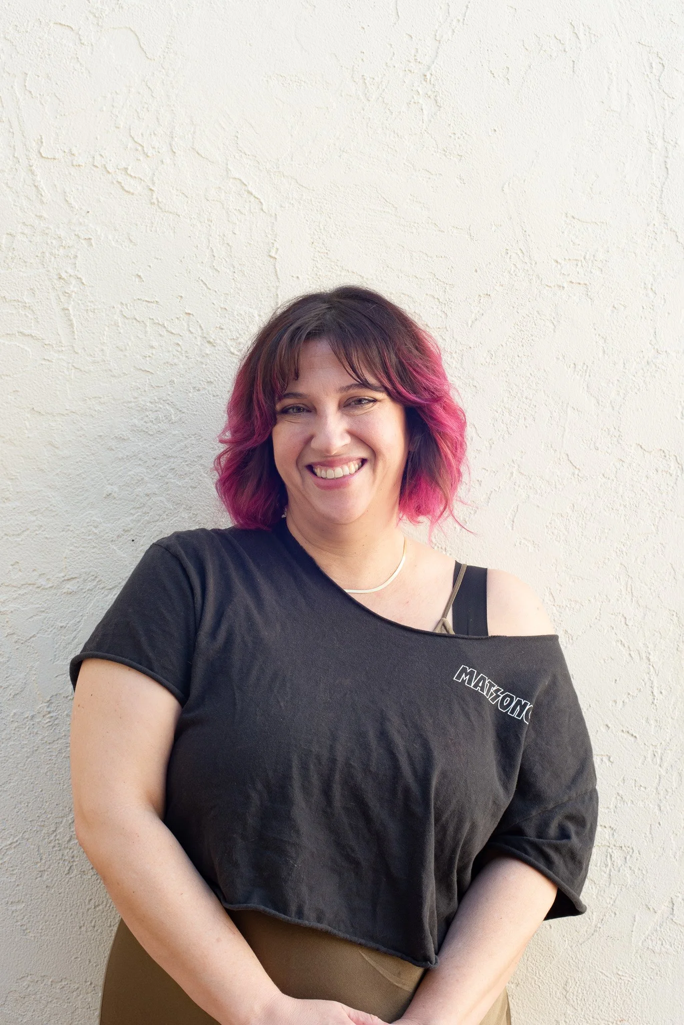 A woman with shoulder-length wavy hair dyed pink and wearing a black off-the-shoulder T-shirt, smiling and standing against a textured white wall.