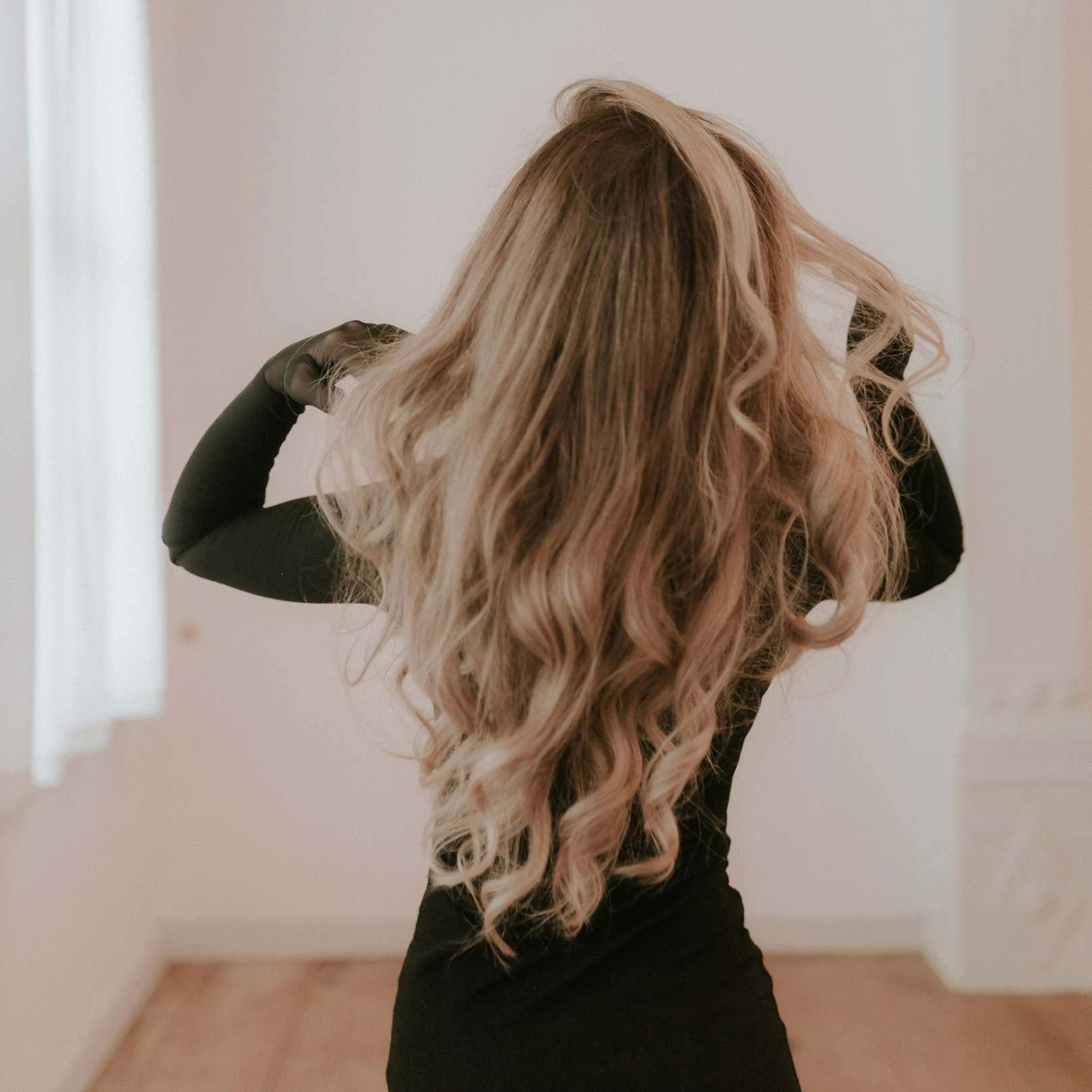 Woman with long, wavy blonde hair adjusting her black dress in a bright room.