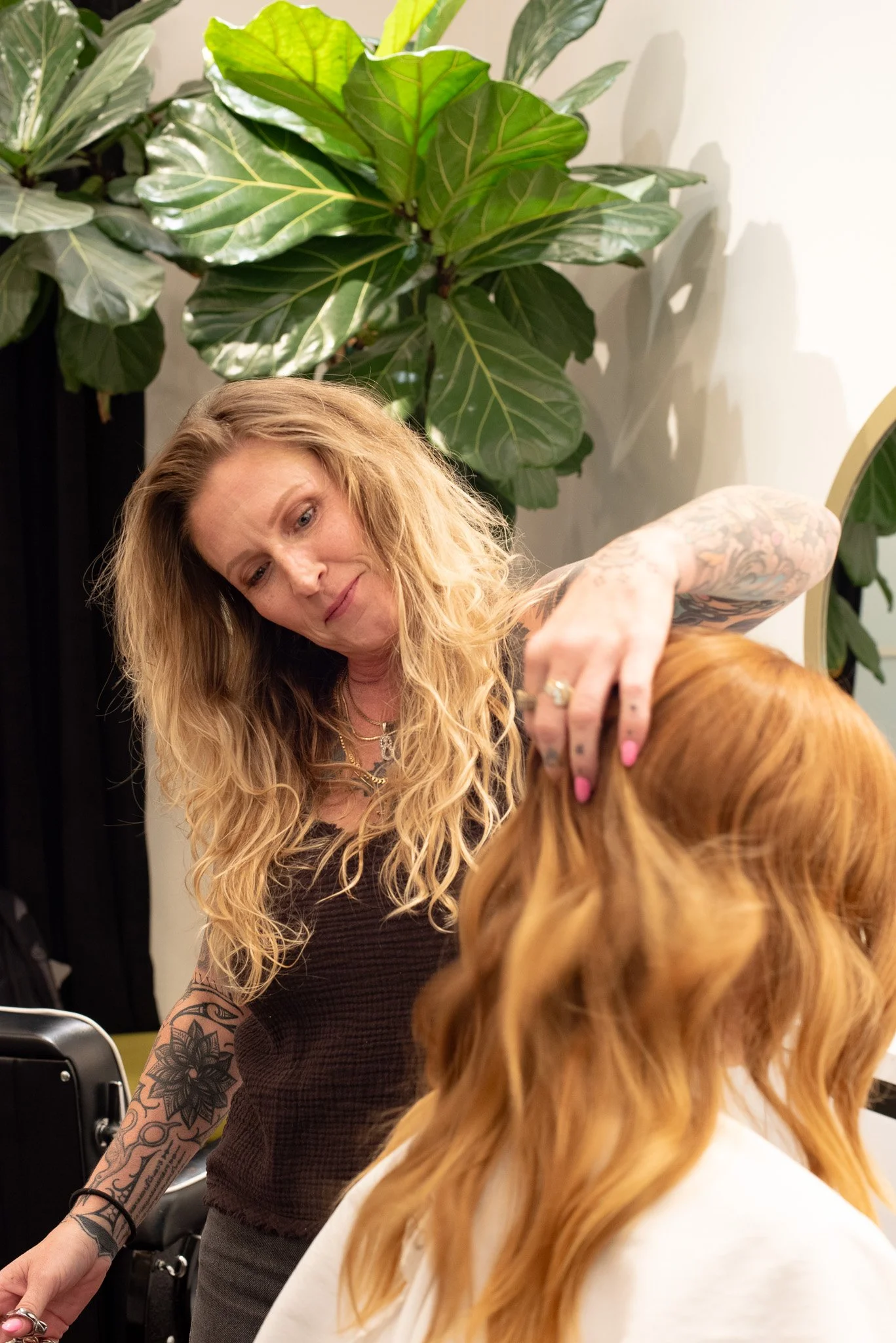 A woman with long wavy blonde hair and tattoos on her arm is styling a woman's red hair, seated in a salon chair. Large green plants are in the background.
