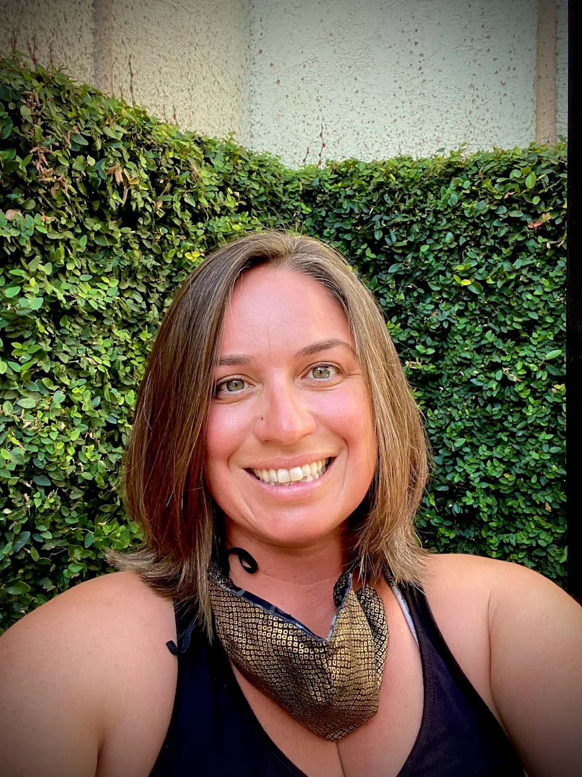 A smiling woman outdoors in front of a green hedge, wearing a face mask around her neck.